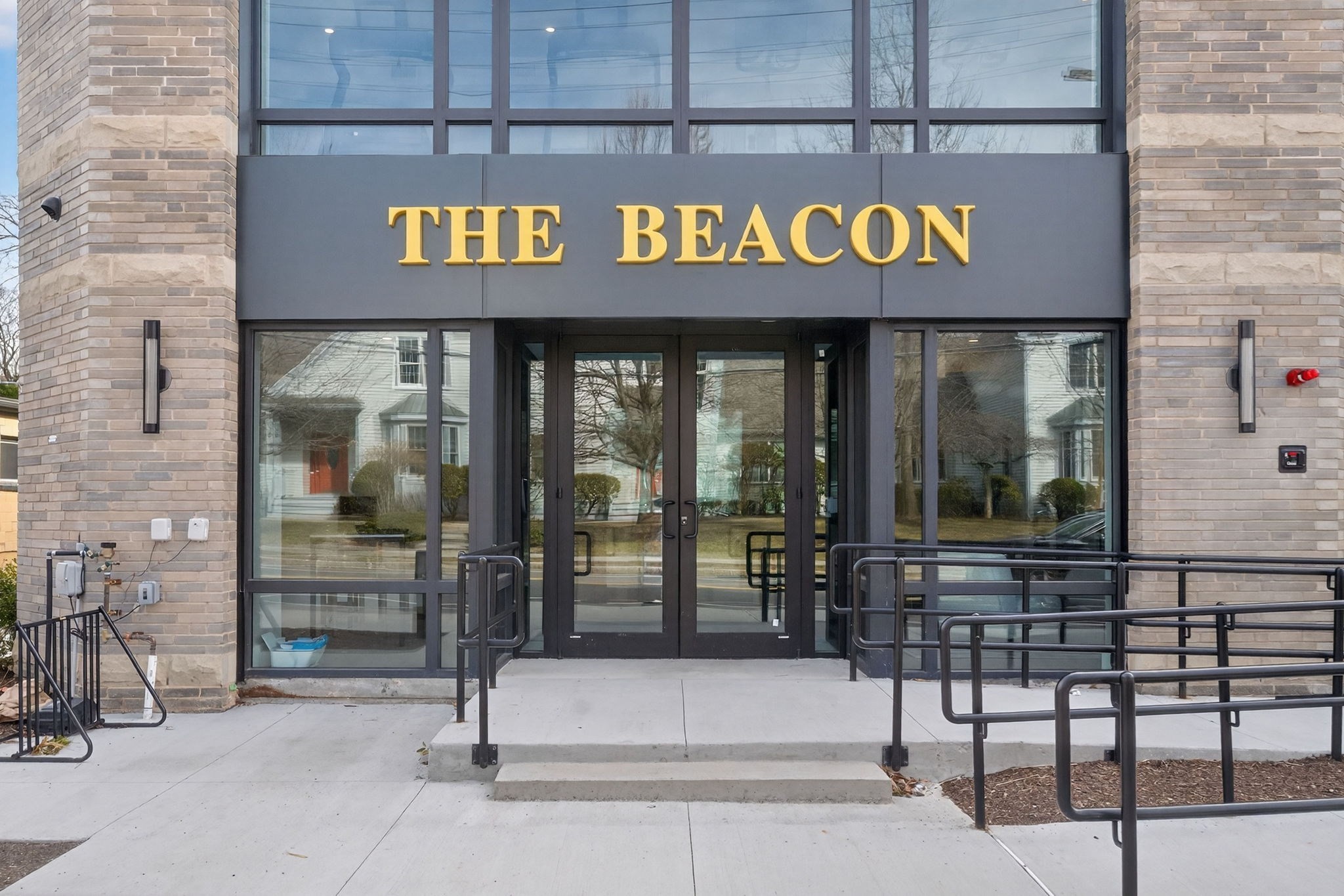 The Beacon condominium entrance with gold lettering, glass doors, brick walls, and accessibility railings.