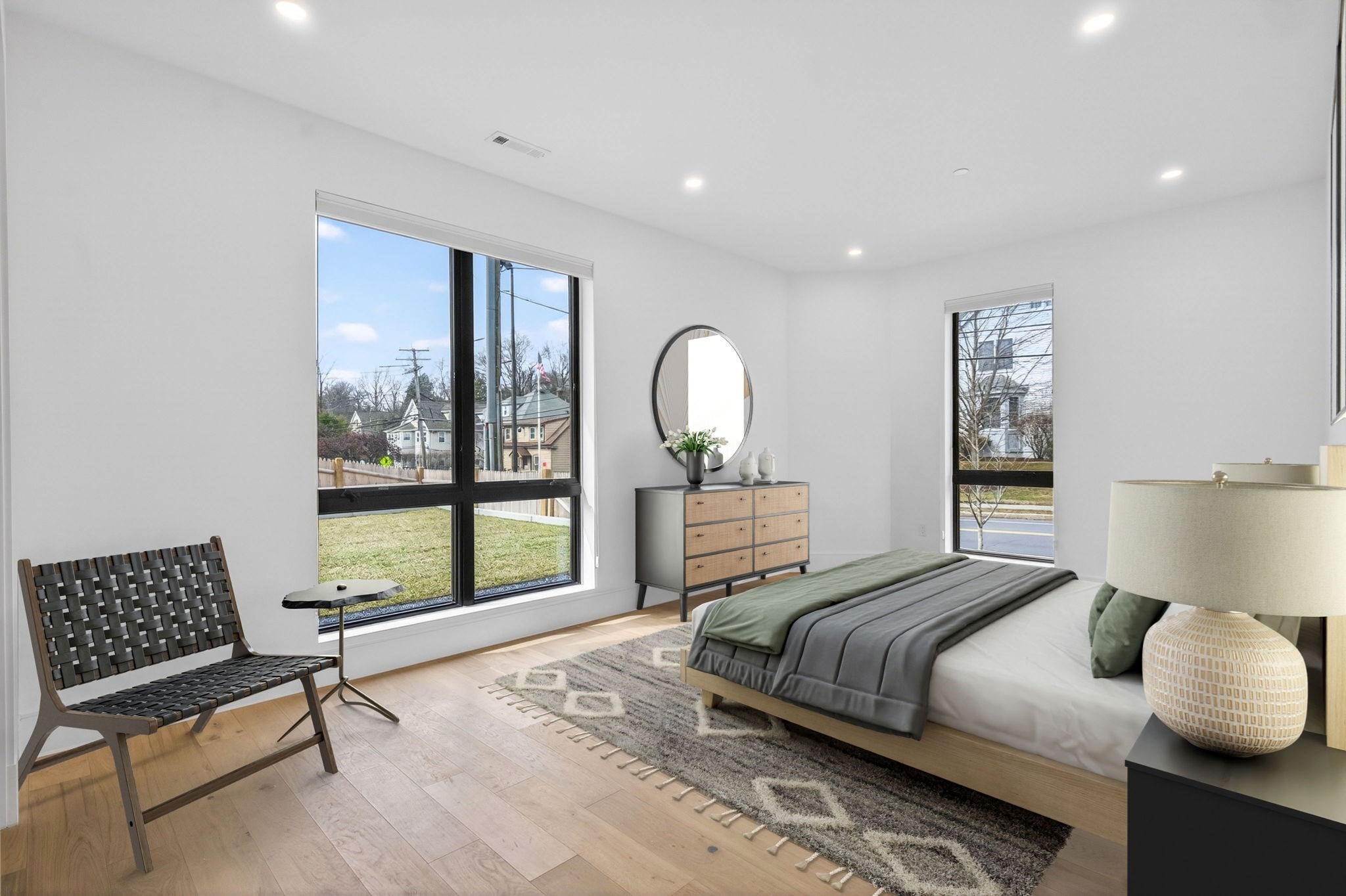 Staged bedroom with gray and sage bedding, a woven chair, and large windows overlooking the neighborhood.