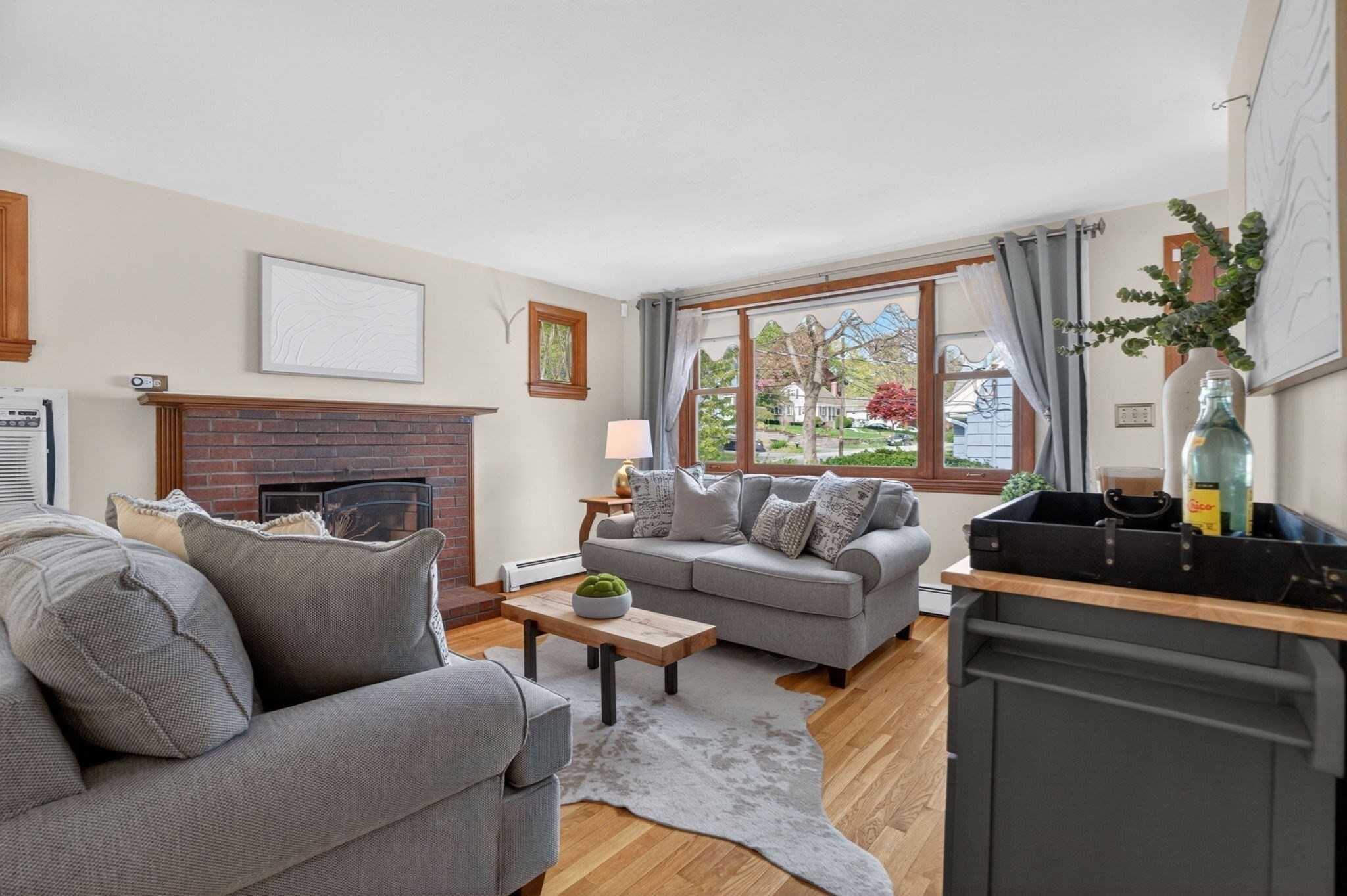 Living room with a brick fireplace, gray sofas, hardwood floors, and a large window overlooking leafy trees.