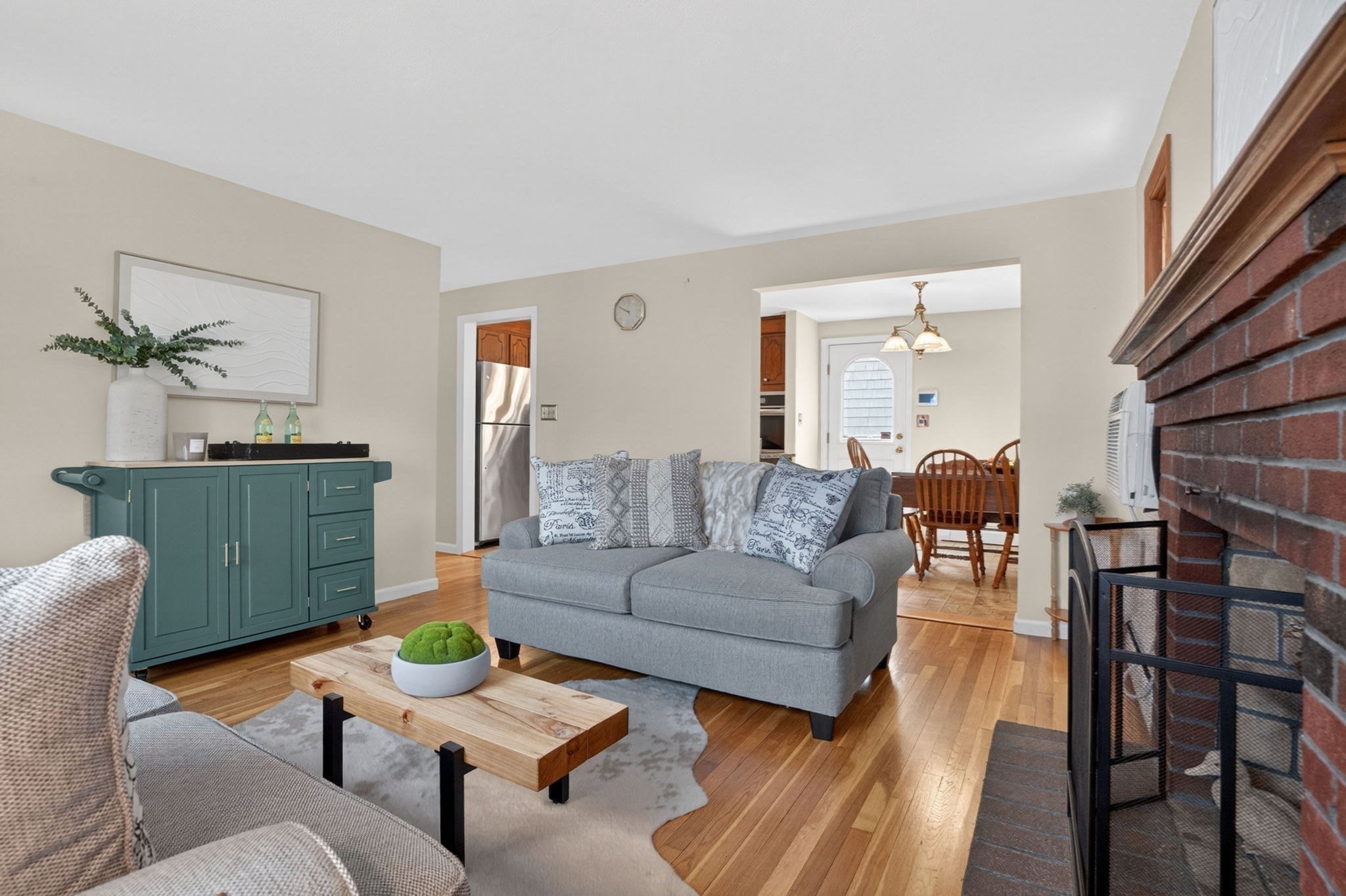 Living room with gray sofa, teal sideboard, hardwood floors, and brick fireplace opening to dining area.