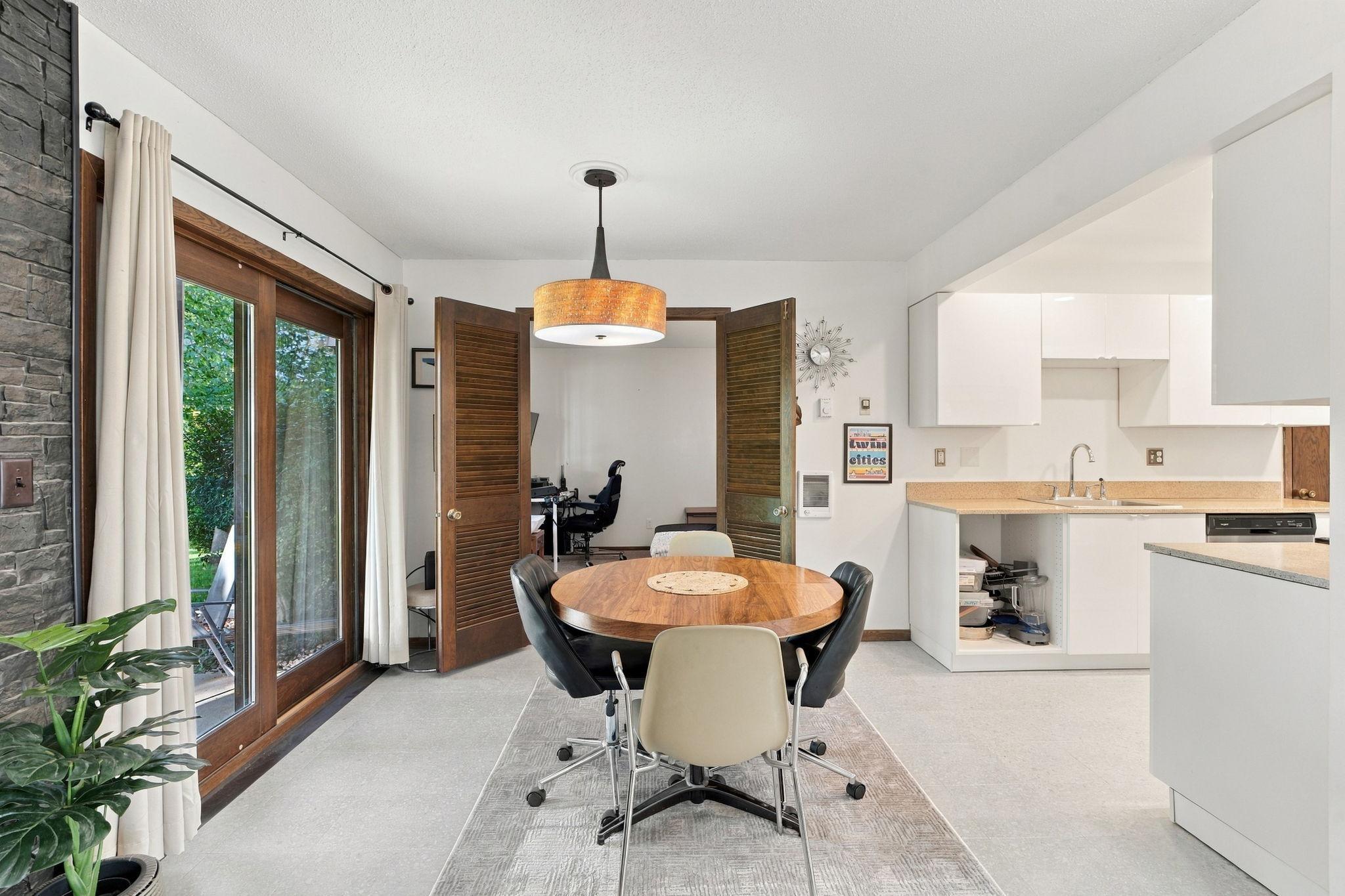 Dining area with a round wooden table beside tall glass patio doors and an adjoining white kitchen.