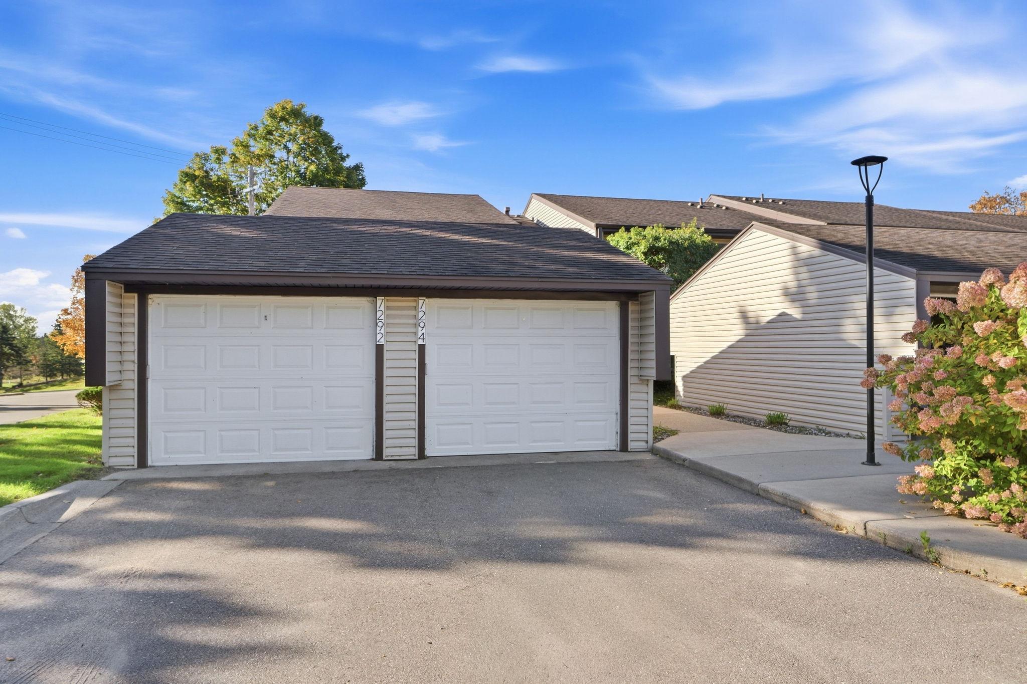 Twin white garage doors marked 7292 and 7294 beside a paved driveway and flowering shrubs.
