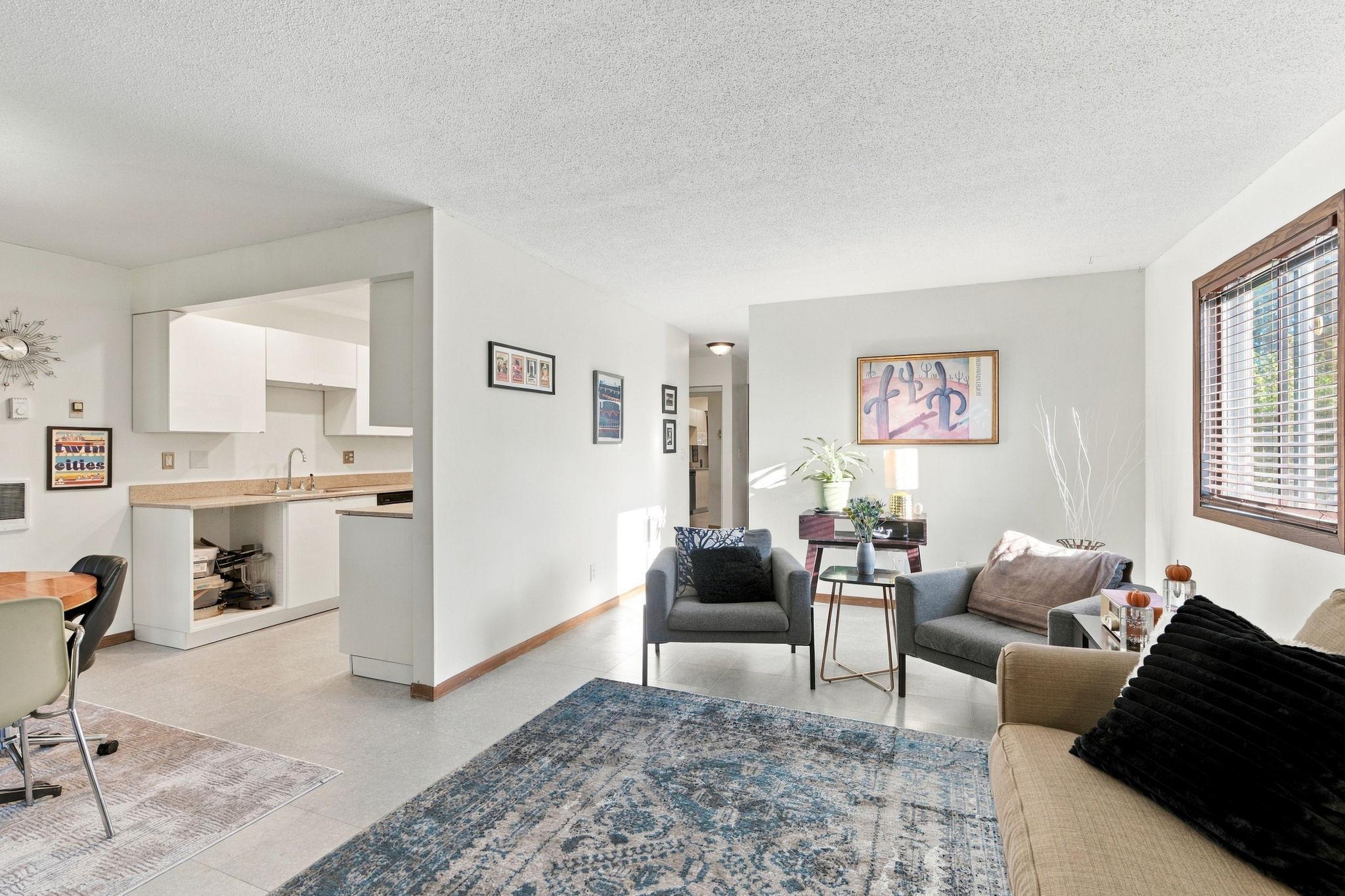 Open-plan living room with gray seating, blue patterned rug, wood-trimmed window, and adjoining white kitchen.