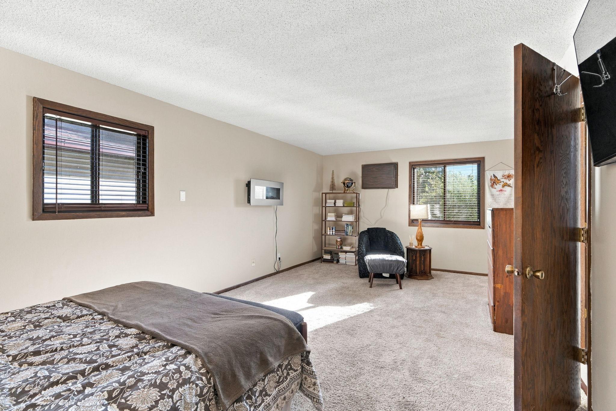 Furnished bedroom with patterned bed, carpet, and a sunny sitting area by the window.