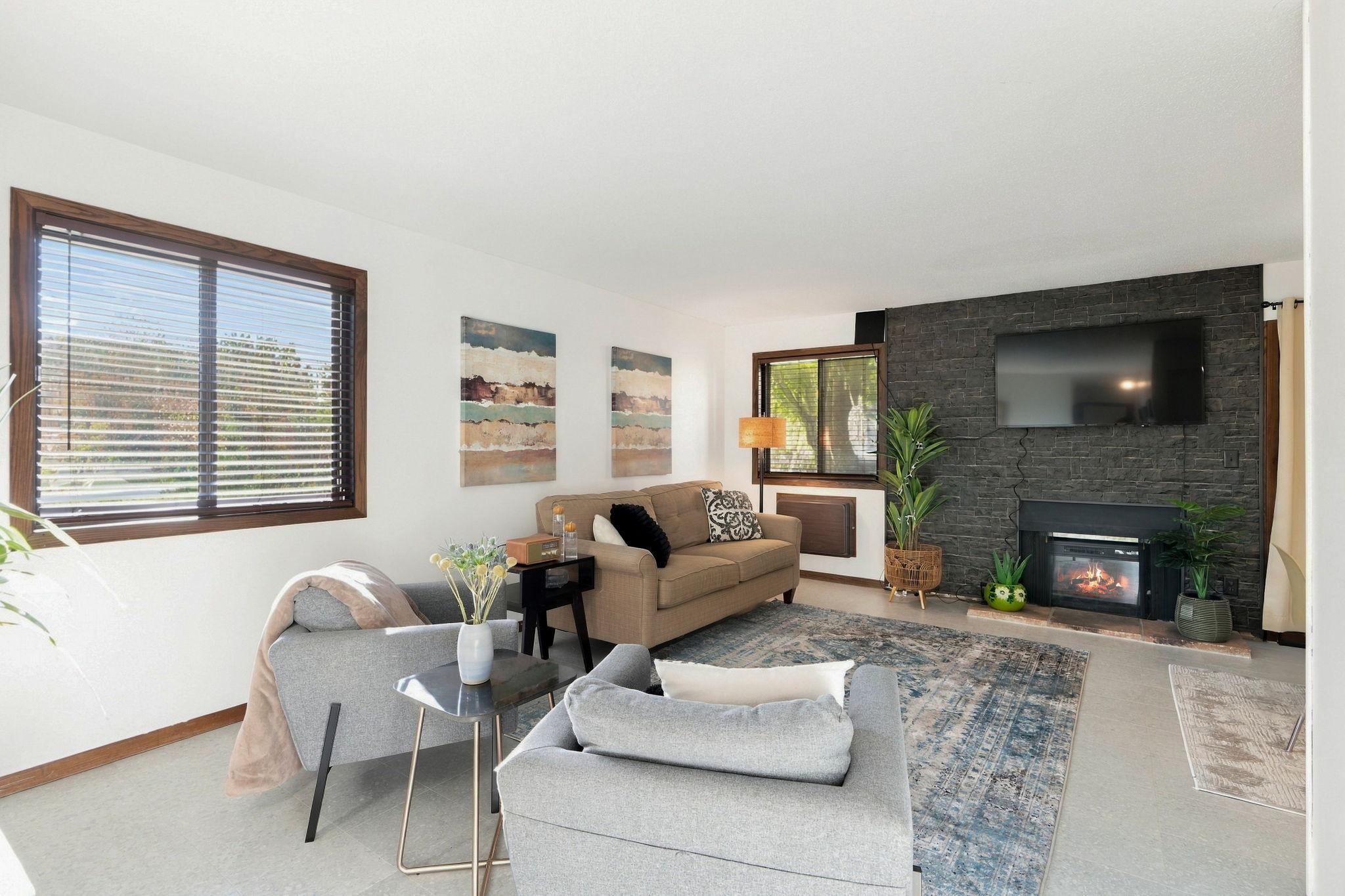 Furnished living room with a dark stone fireplace wall, mounted television, and large wood-trimmed windows.