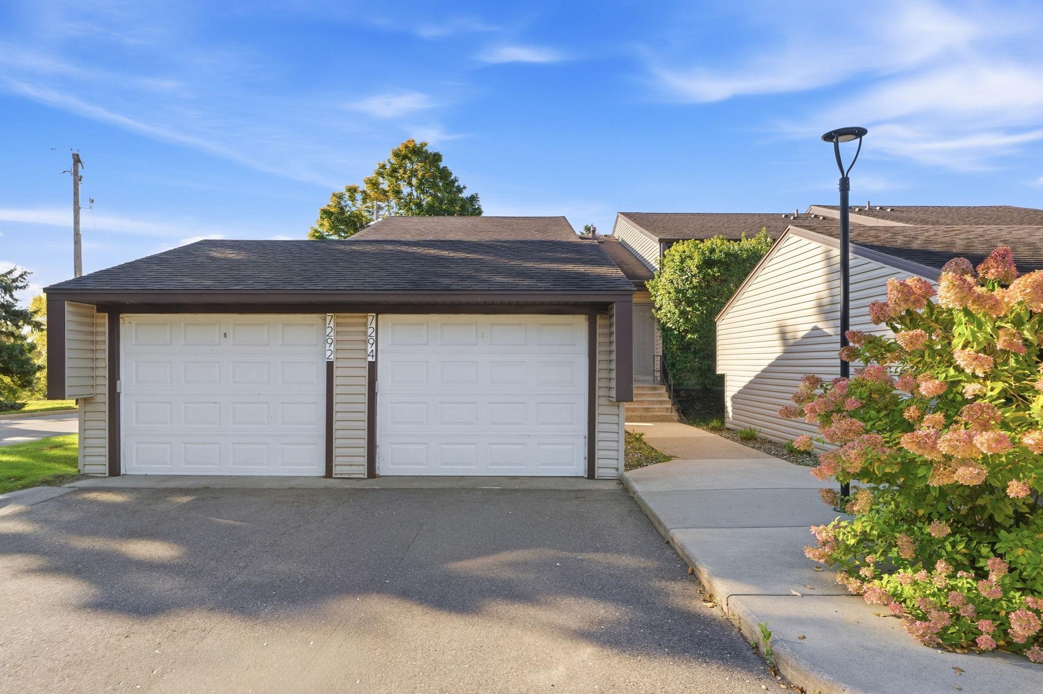 Two-car garage with white paneled doors, dark trim, and flowering shrubs along the driveway.