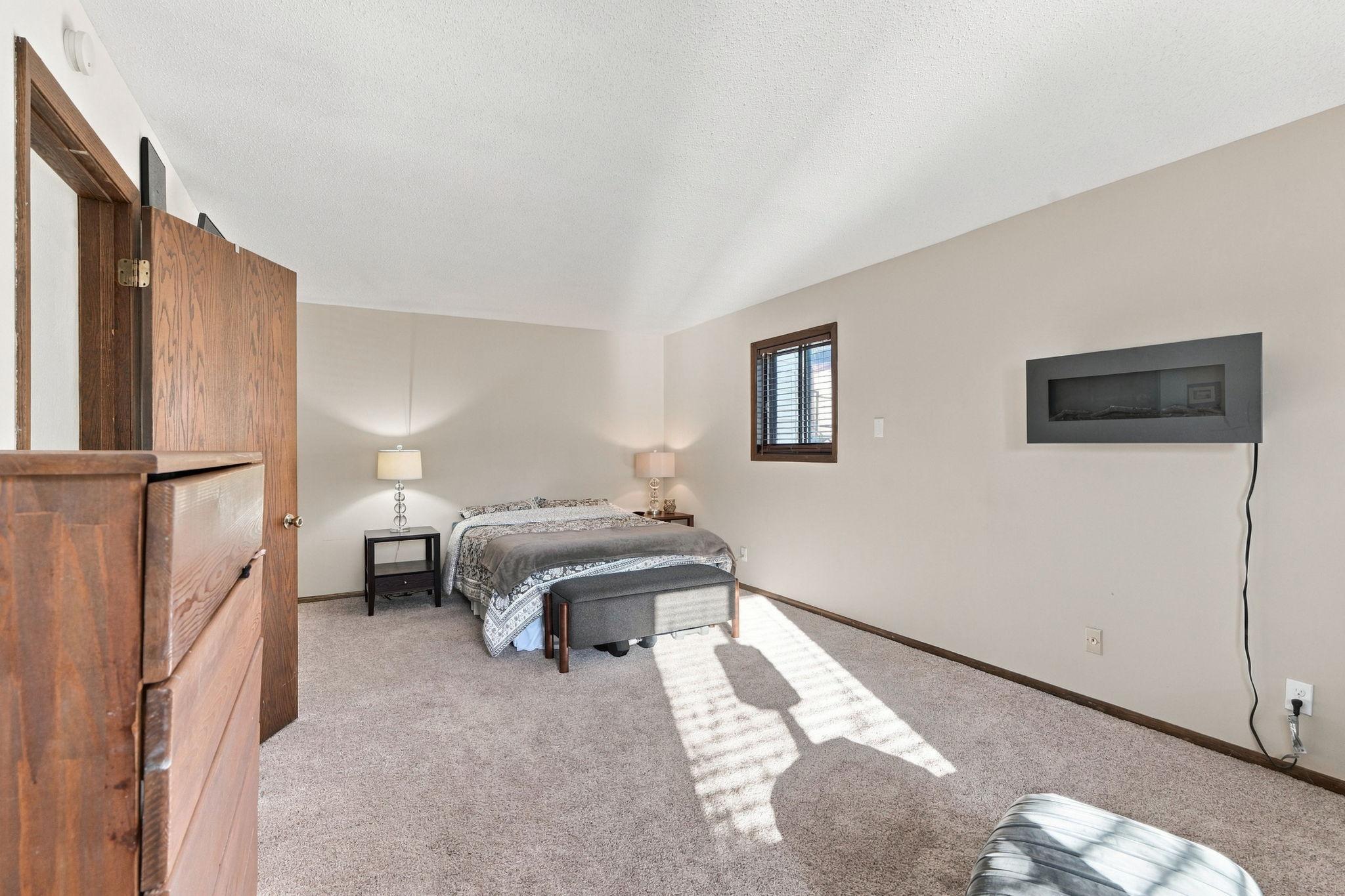 Furnished bedroom with a bed, carpeted floor, and modern wall-mounted fireplace.