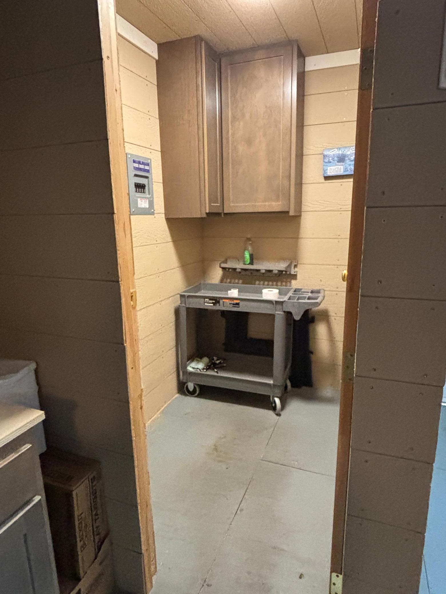 Utility room with tan paneled walls, an electrical panel, overhead cabinets, and a rolling workbench.