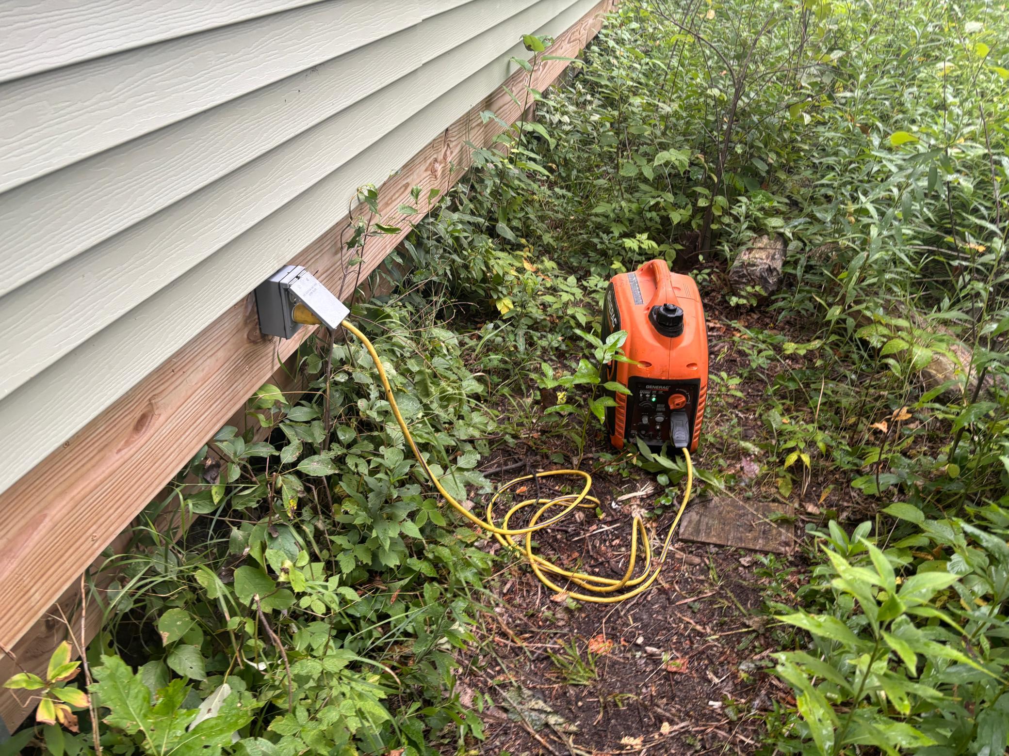 Orange portable generator plugged into an outdoor outlet beside the house, surrounded by overgrown vegetation.