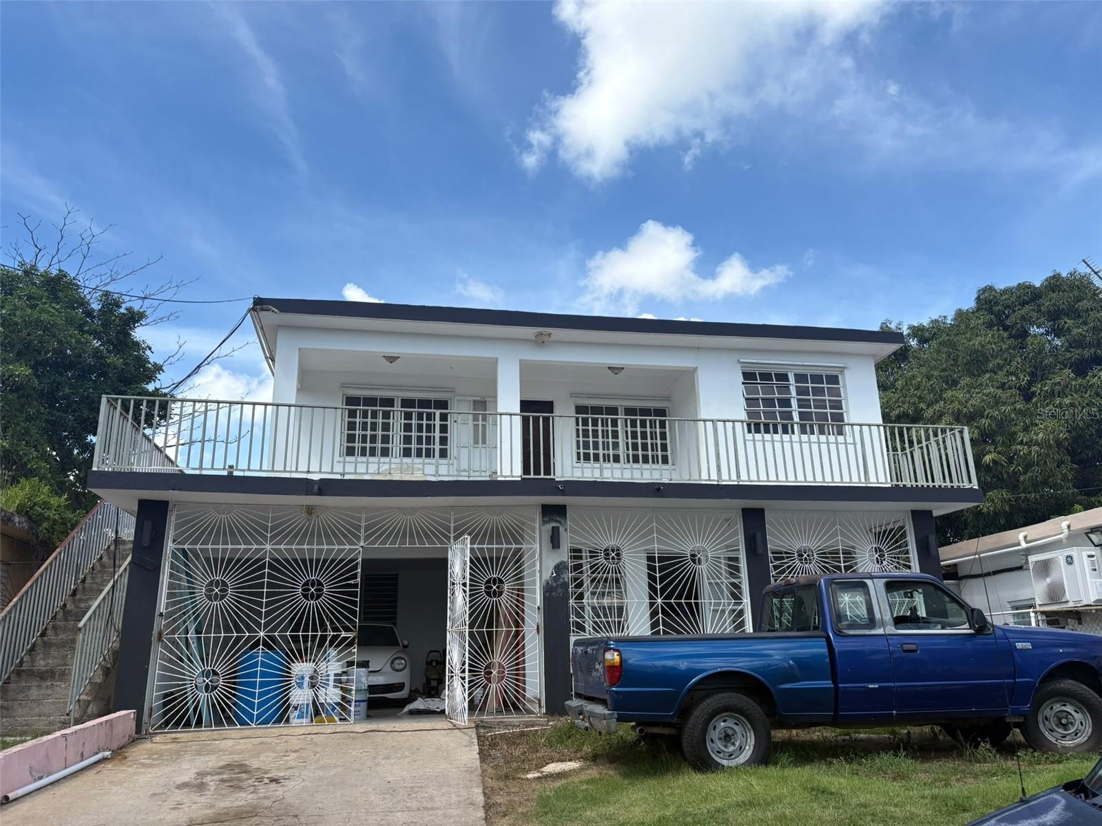 Two-story white residence with a long balcony, decorative metal gates, and a blue pickup in the driveway.