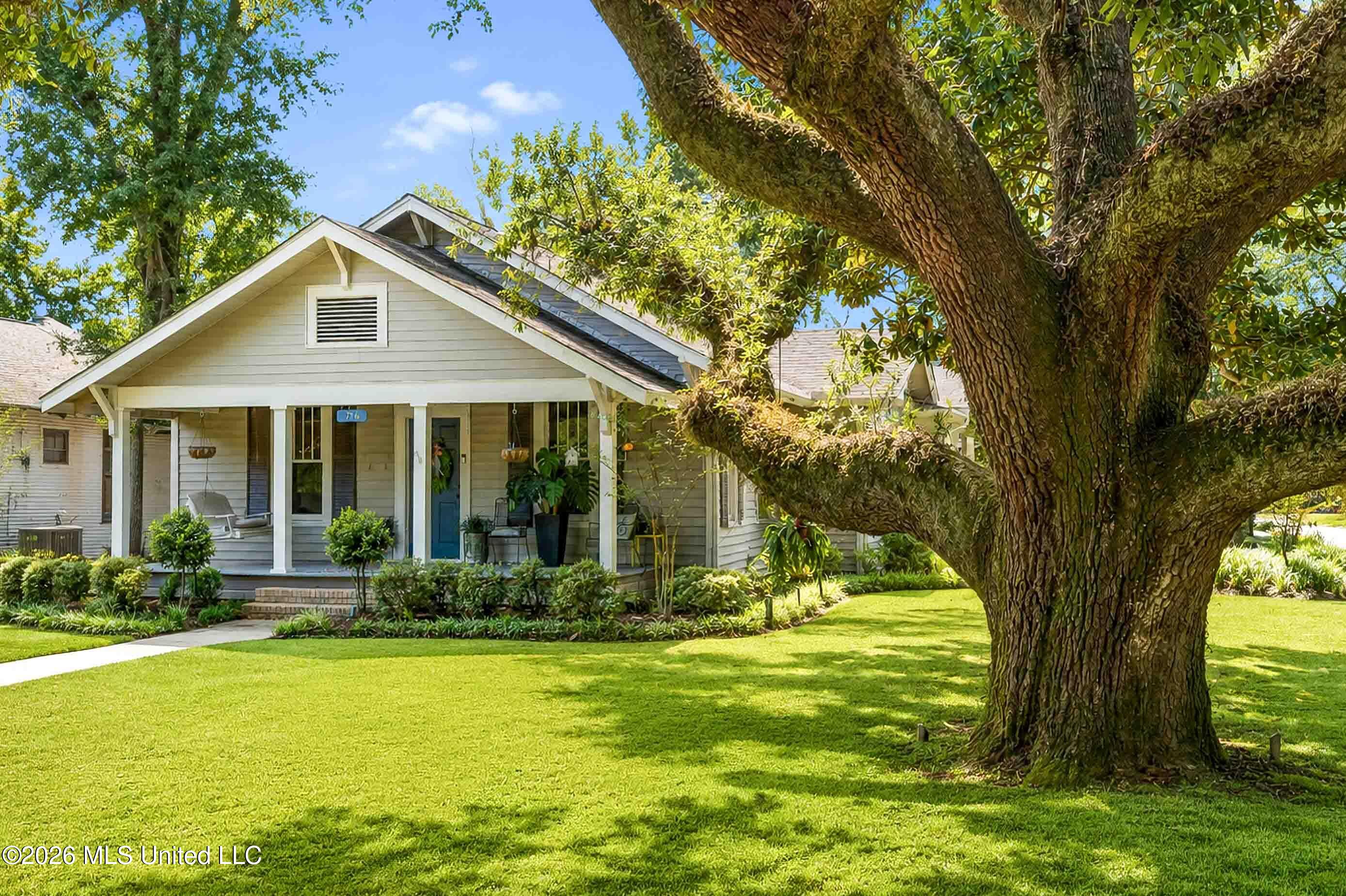 Gray gabled house with a covered porch and blue door, framed by a large tree and landscaped lawn.