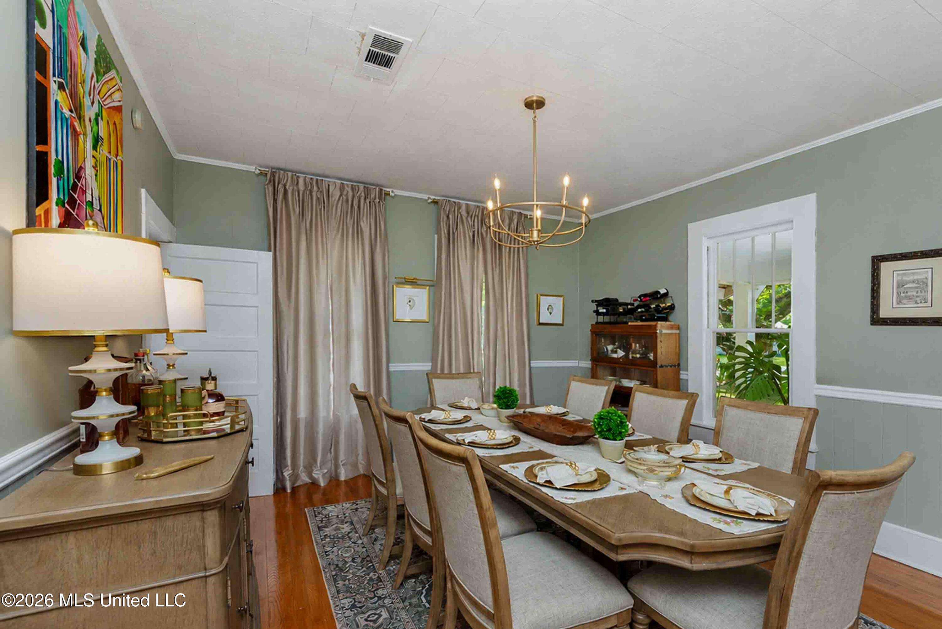 Staged dining room with a set table, upholstered chairs, brass chandelier, and tall curtained windows.