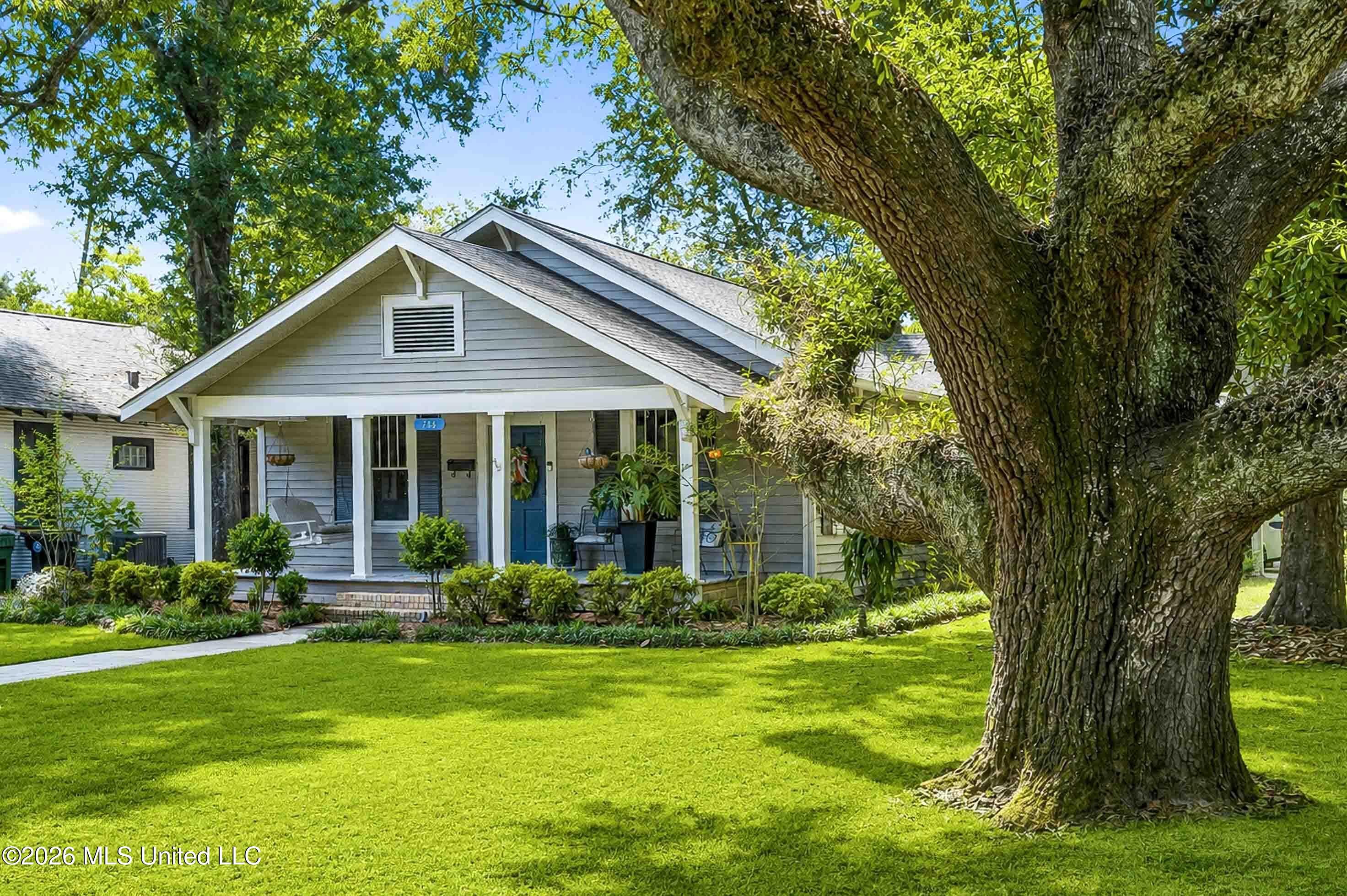 Gray house with a blue front door and covered porch, framed by a large shade tree and green lawn.