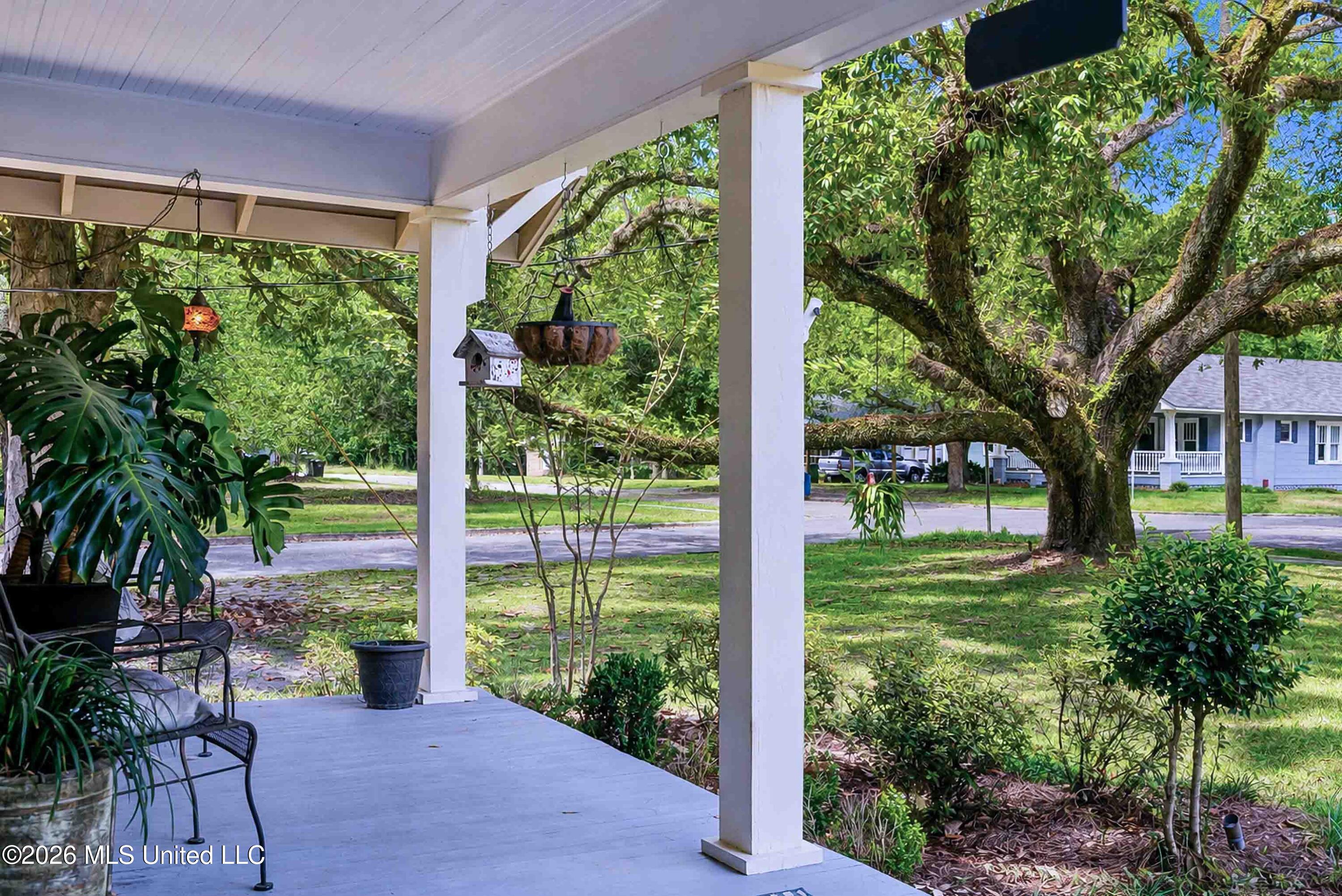 Covered porch with metal chairs and potted plants overlooking a leafy yard and a large tree.