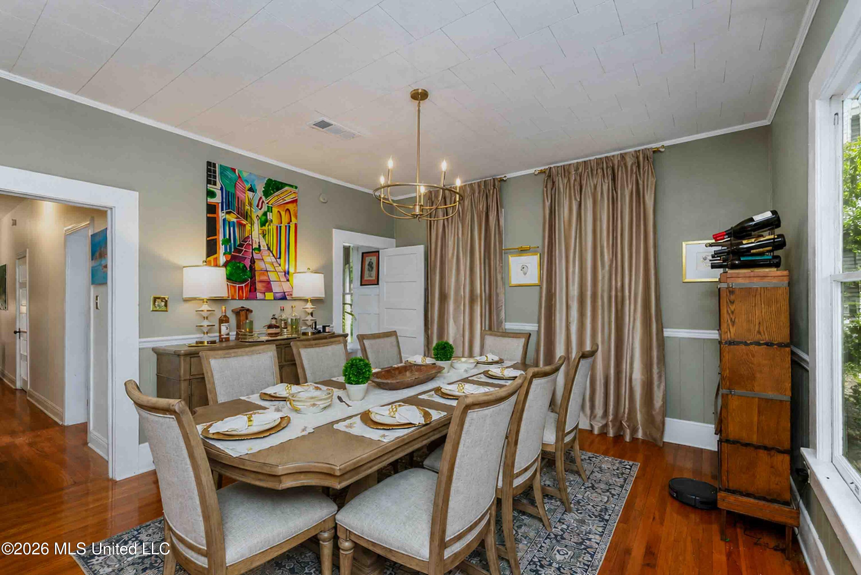 Dining room with a set table, upholstered chairs, brass chandelier, and tall curtained windows.