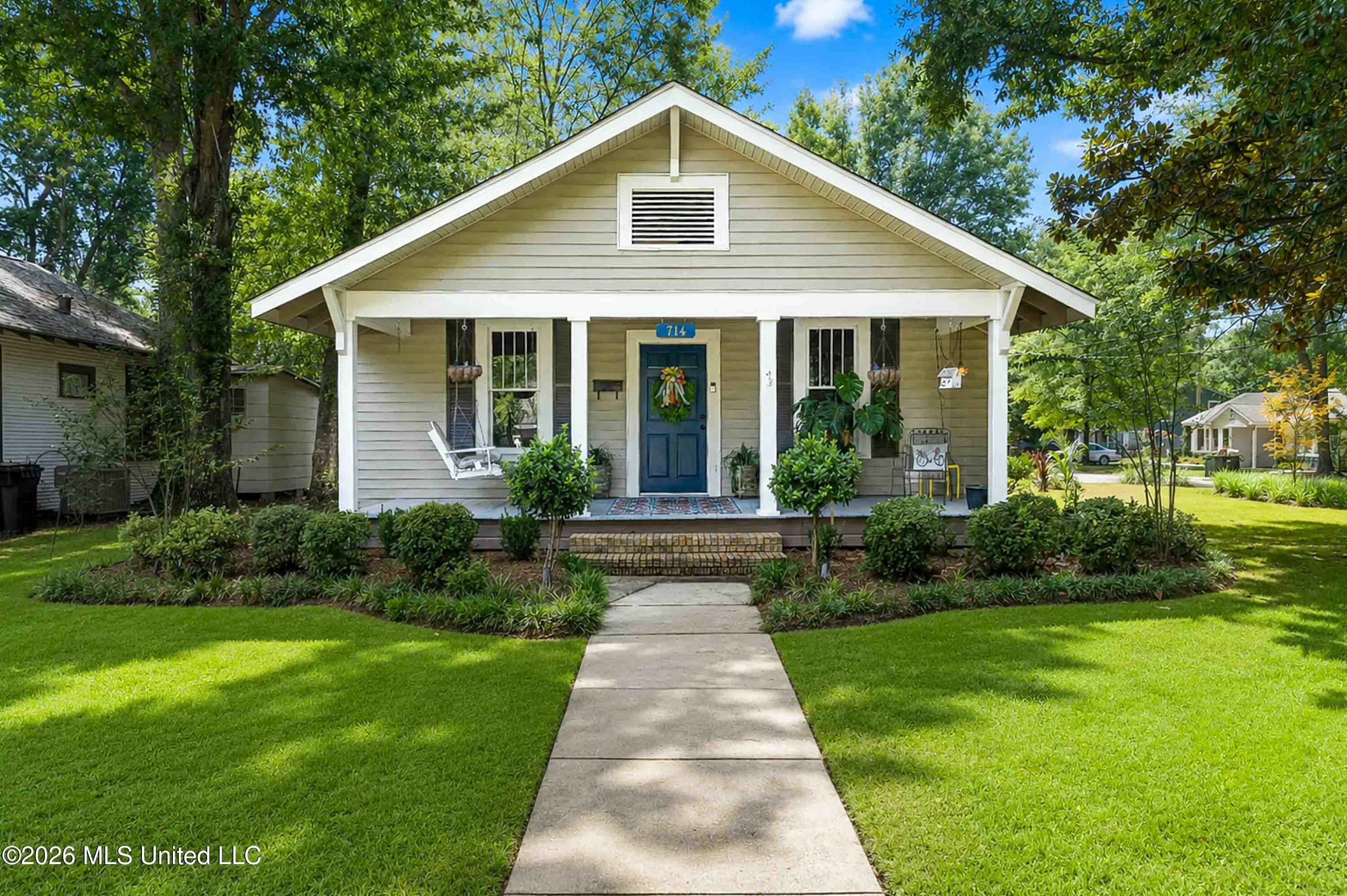 Front of a light-colored house with a blue door, covered porch, and lush landscaping.