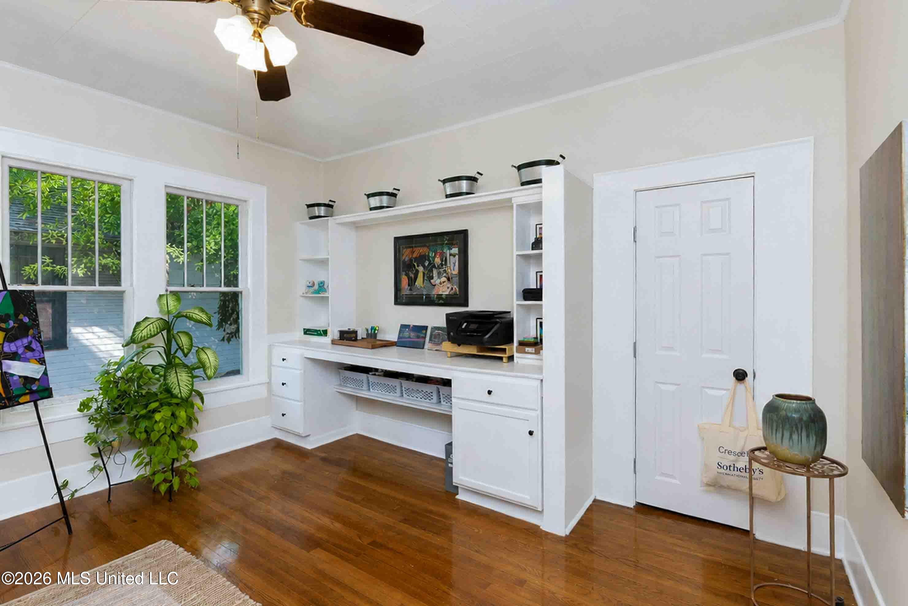 Home office with built-in white desk and shelves, two windows, and polished hardwood floors.