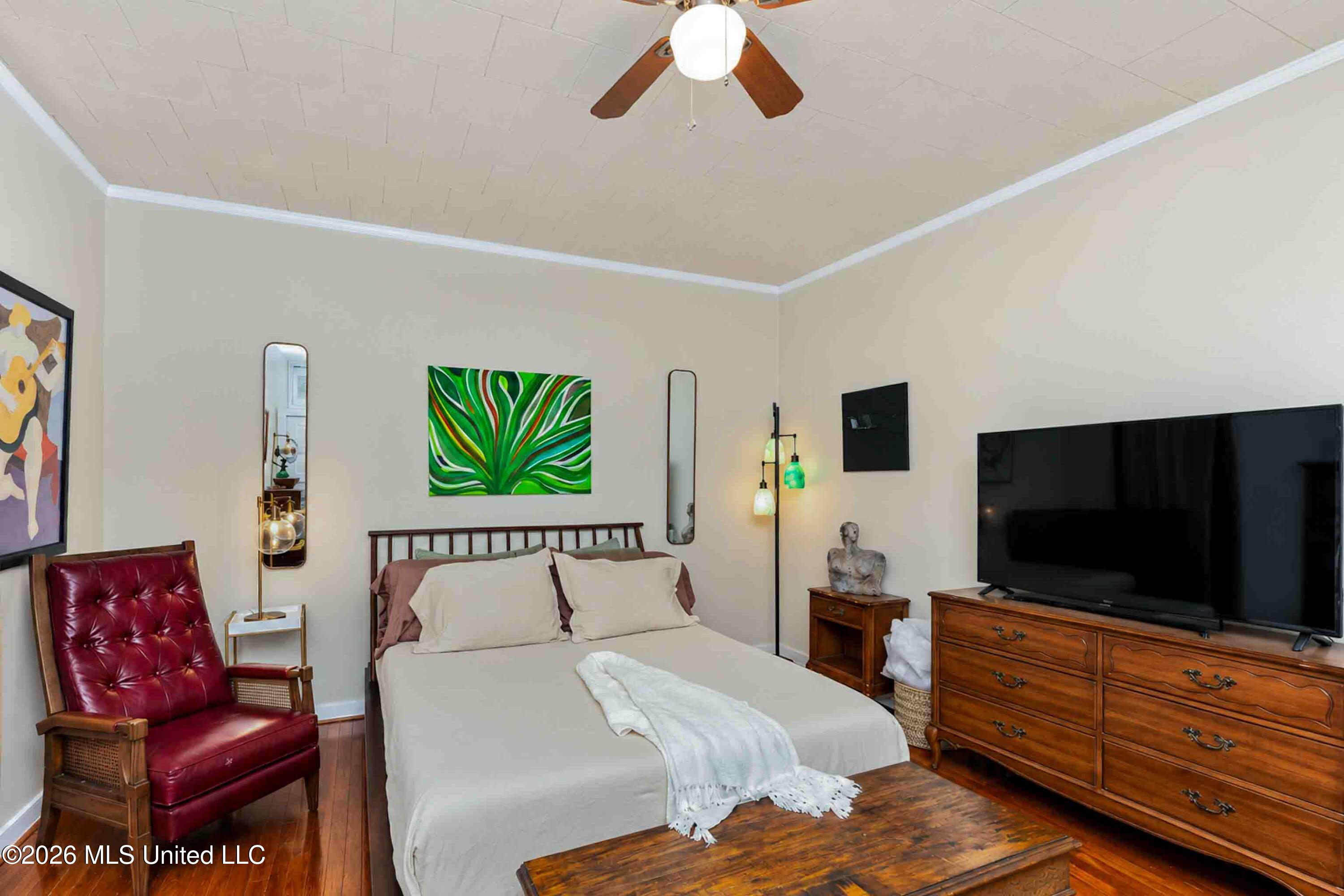 Furnished bedroom with a central bed, hardwood floors, red leather chair, and large television.