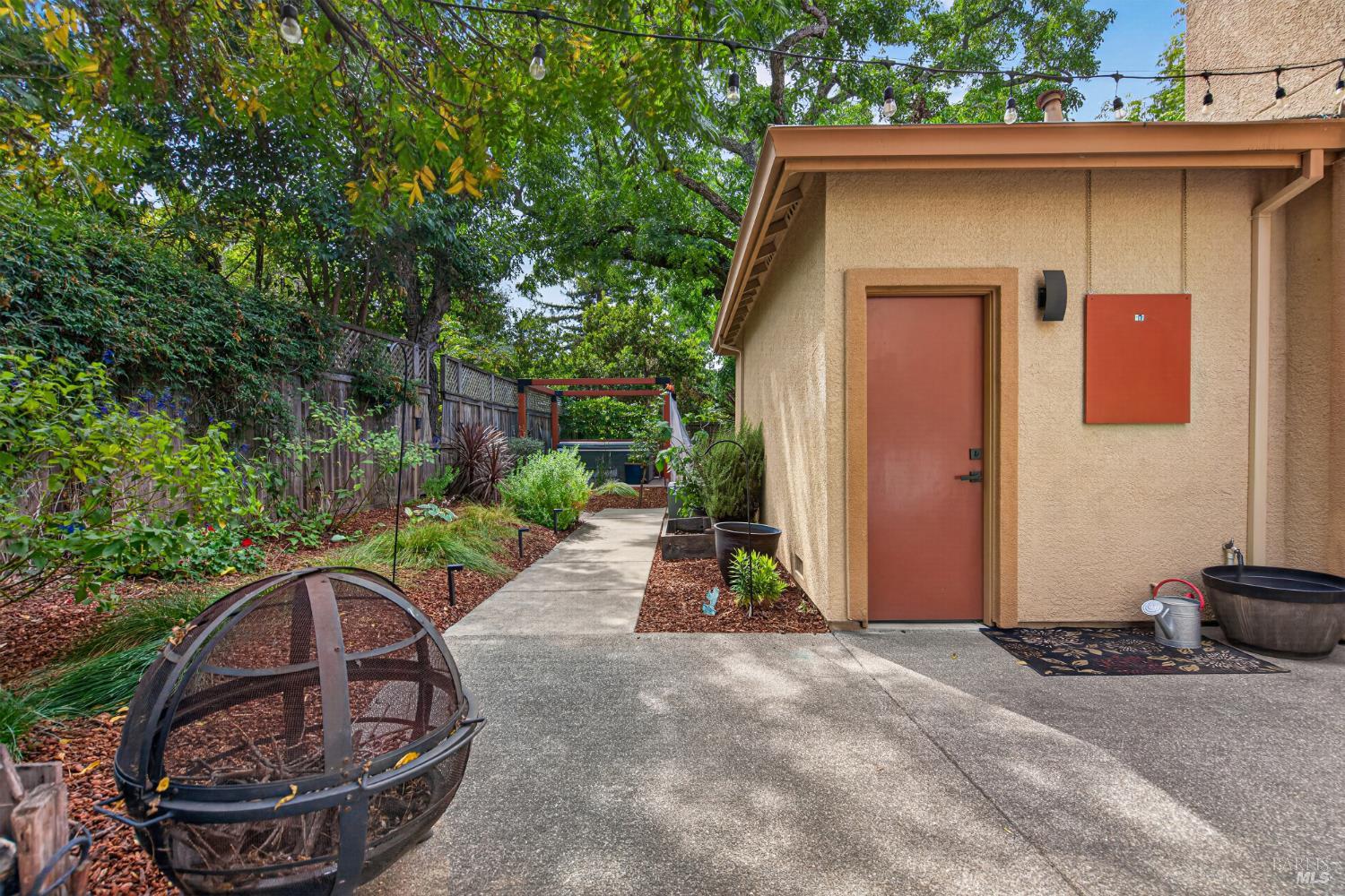 Landscaped backyard walkway beside a tan stucco outbuilding, with overhead string lights and a metal fire pit.