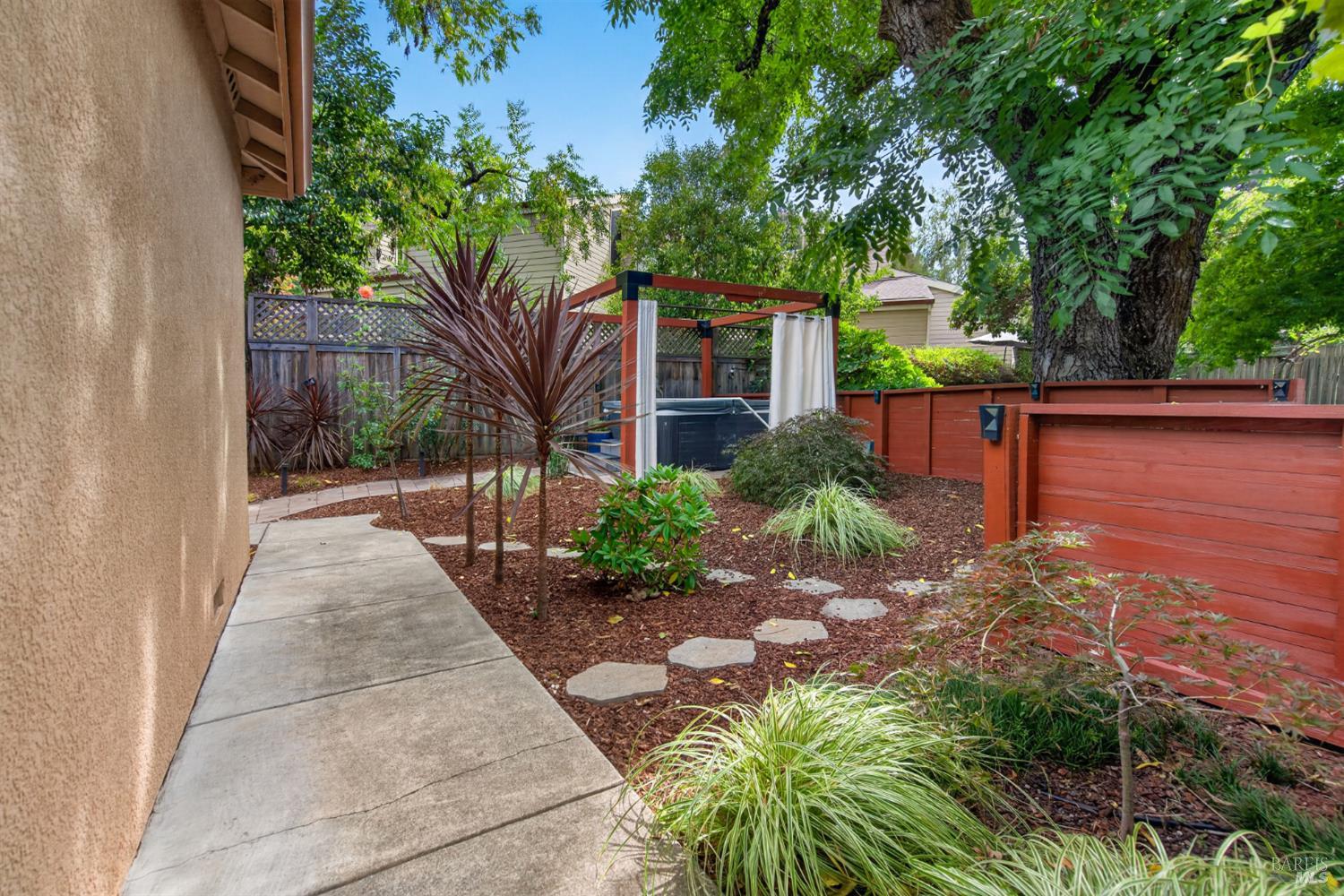Landscaped backyard with a stepping-stone path, red fencing, leafy trees, and a curtained hot tub shelter.