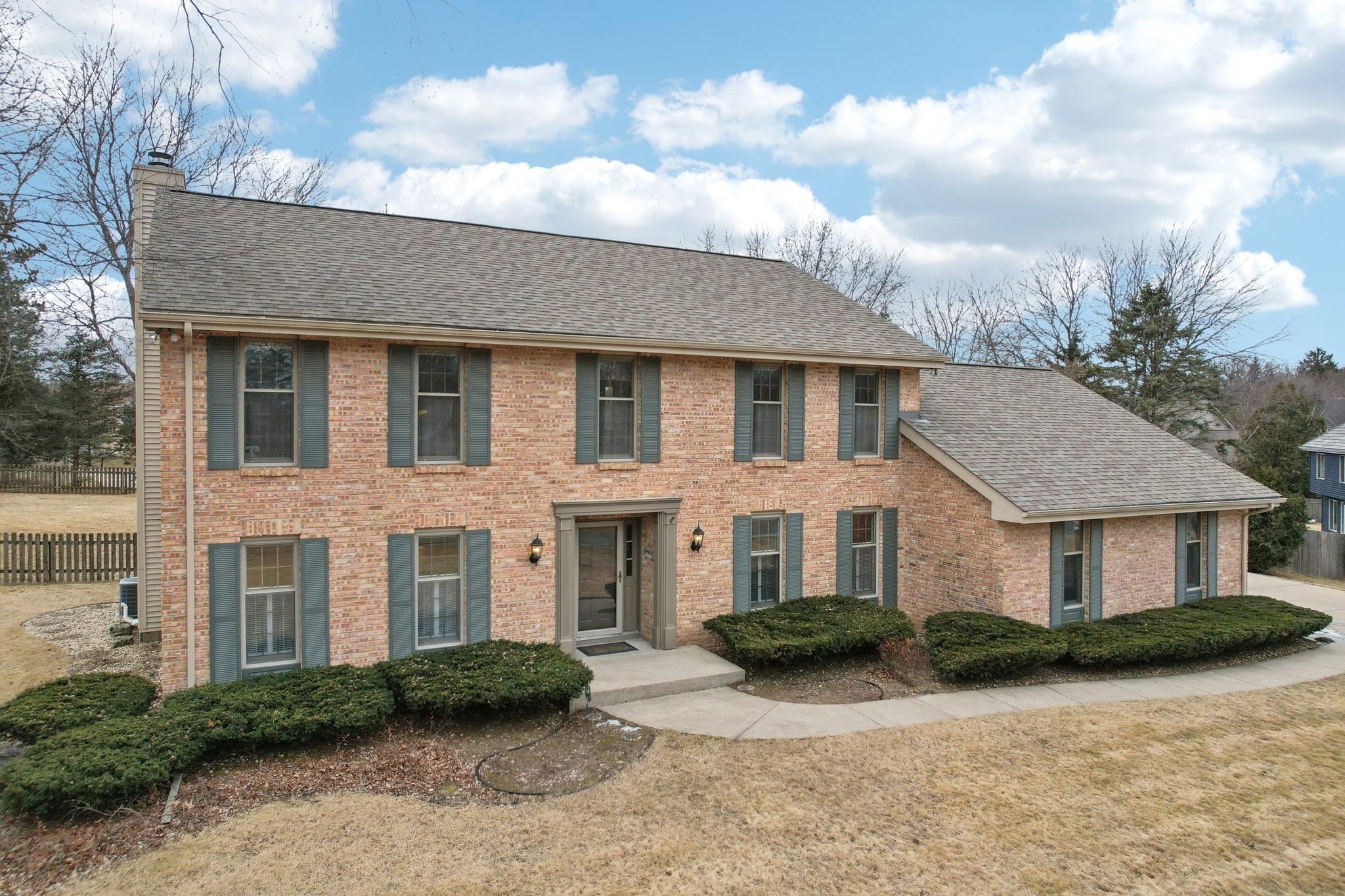 Two-story brick house with gray shutters, an attached side wing, and trimmed front shrubs.