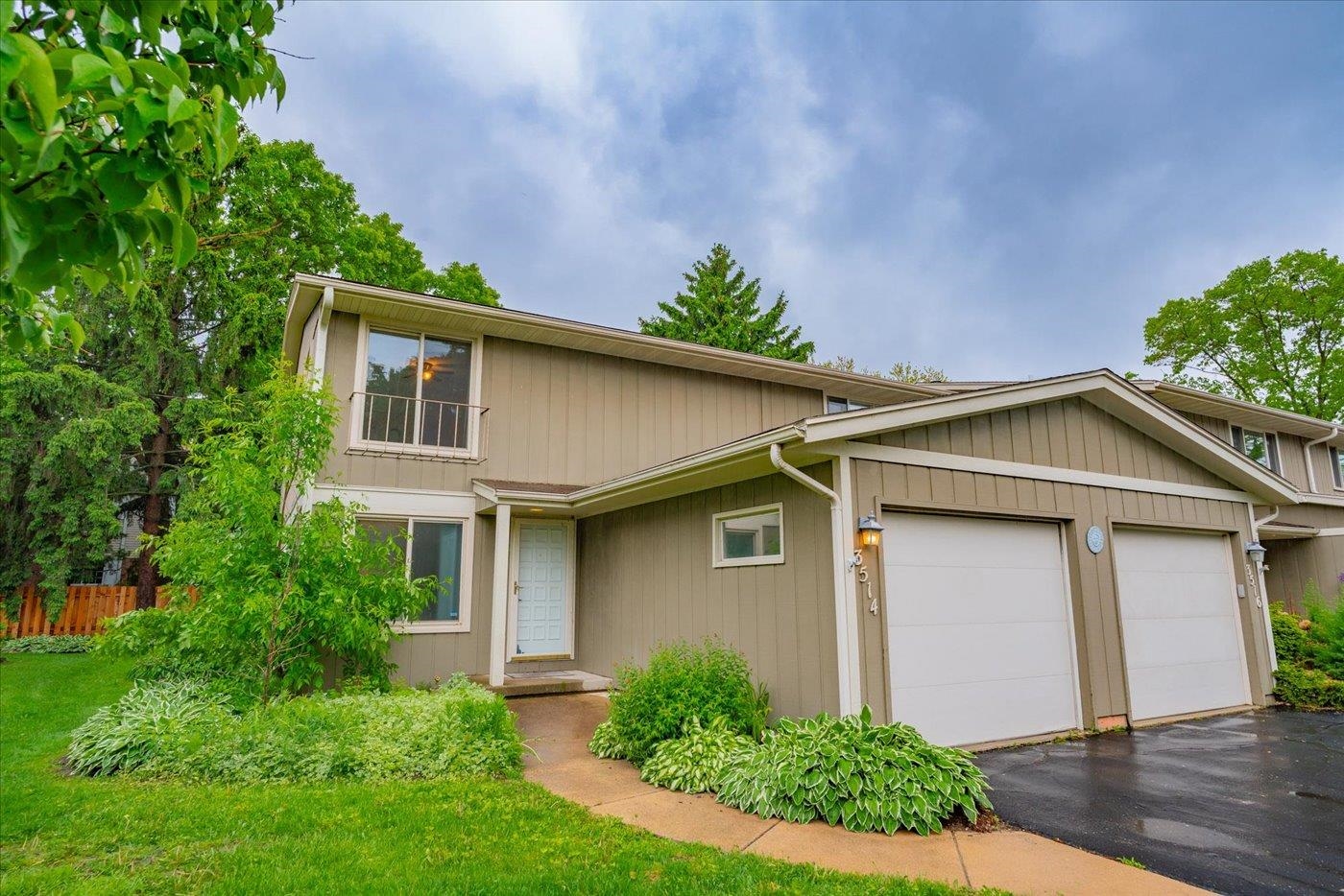 Two-story beige home with two garage doors, an upper balcony, and leafy landscaping.