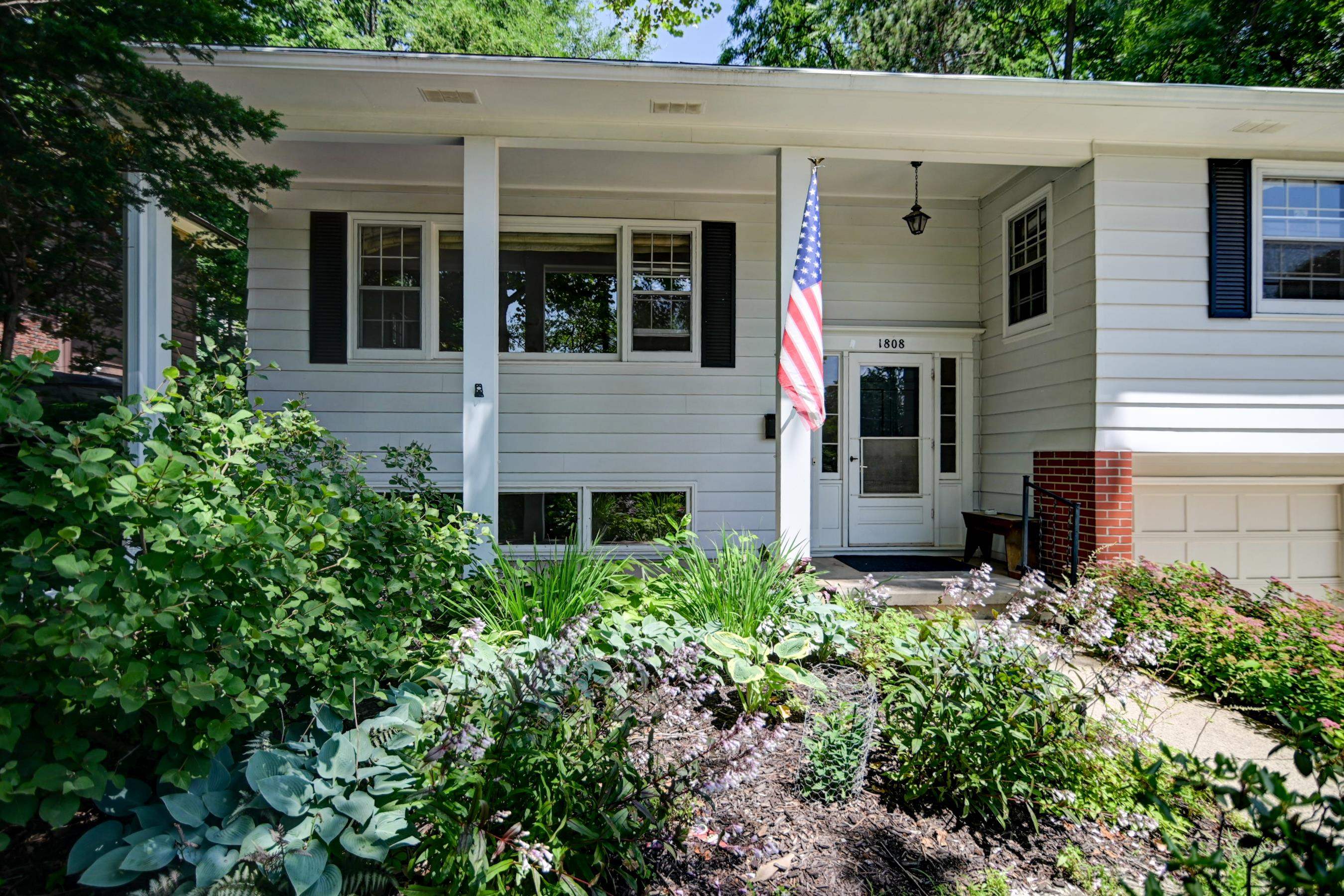 White-sided house with a covered porch, American flag, attached garage, and lush front garden.
