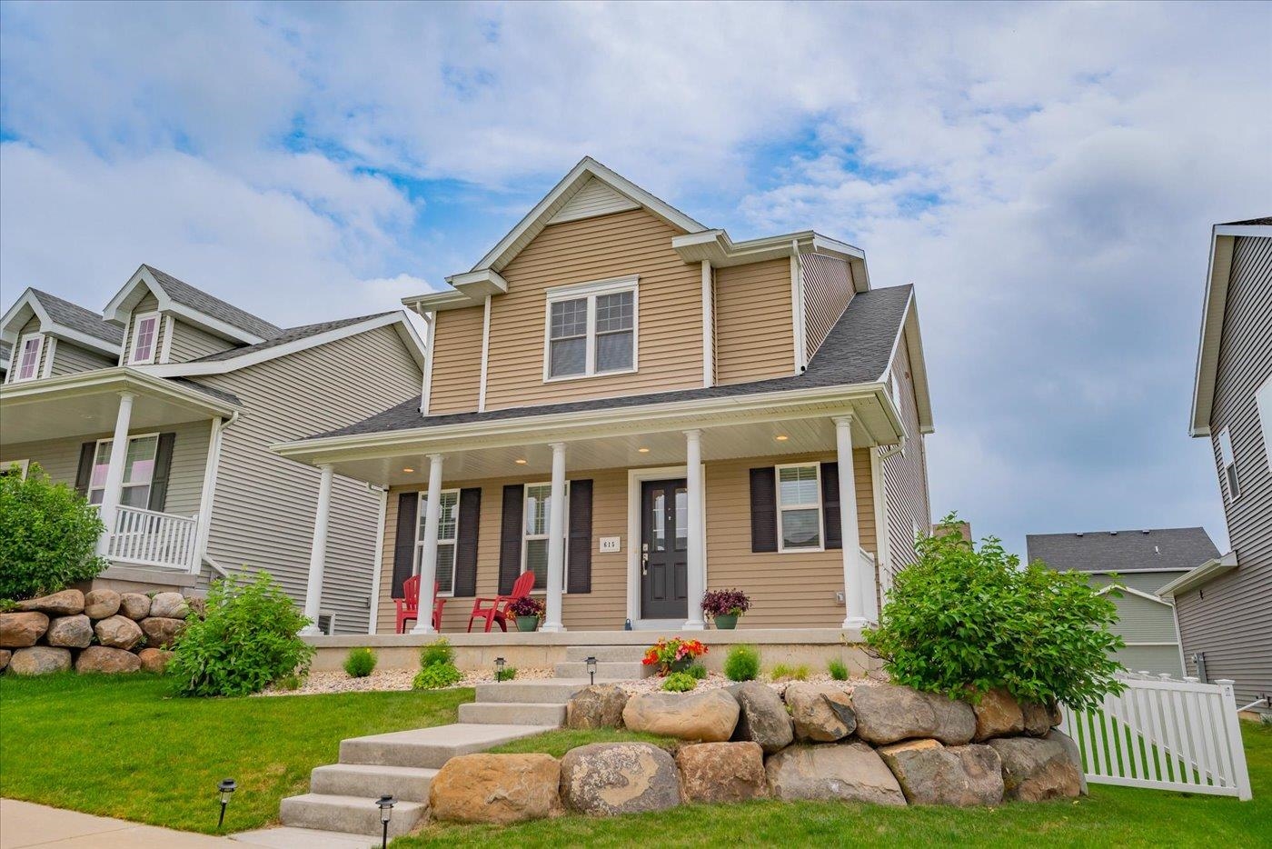 Two-story tan home with a covered porch, red chairs, and a stone retaining wall.