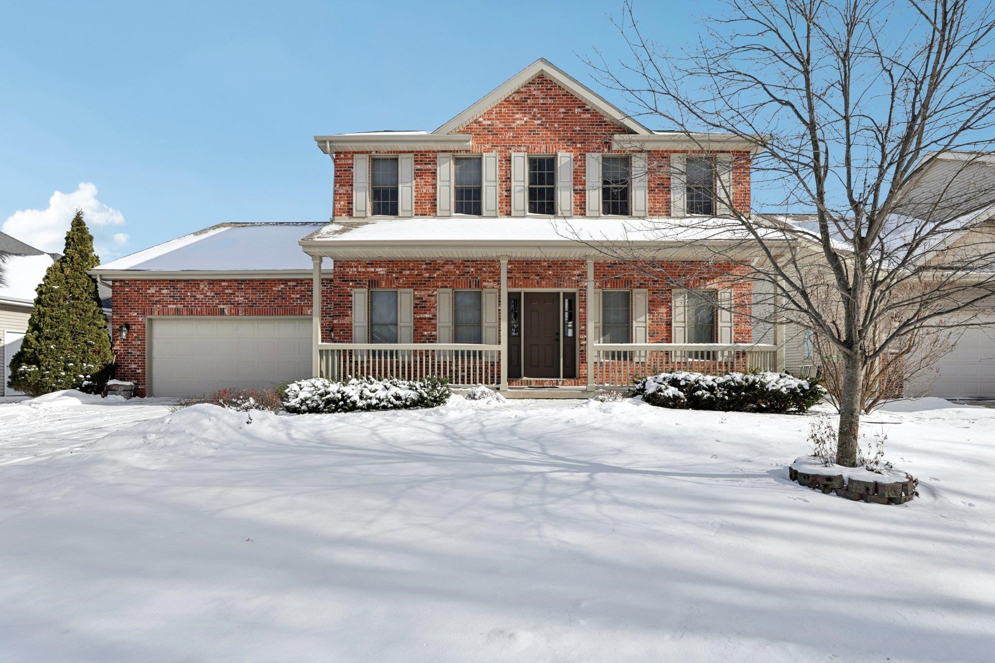 Two-story red brick house with a covered porch and attached garage, surrounded by snow.