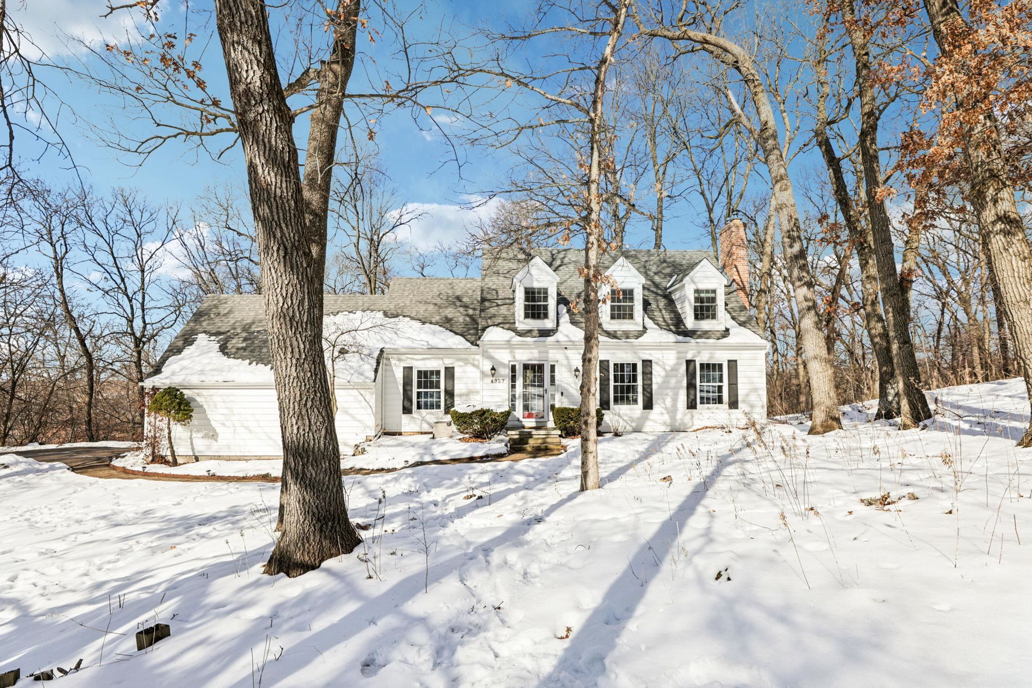 White two-story house with dormers and black shutters, surrounded by snow and tall bare trees.