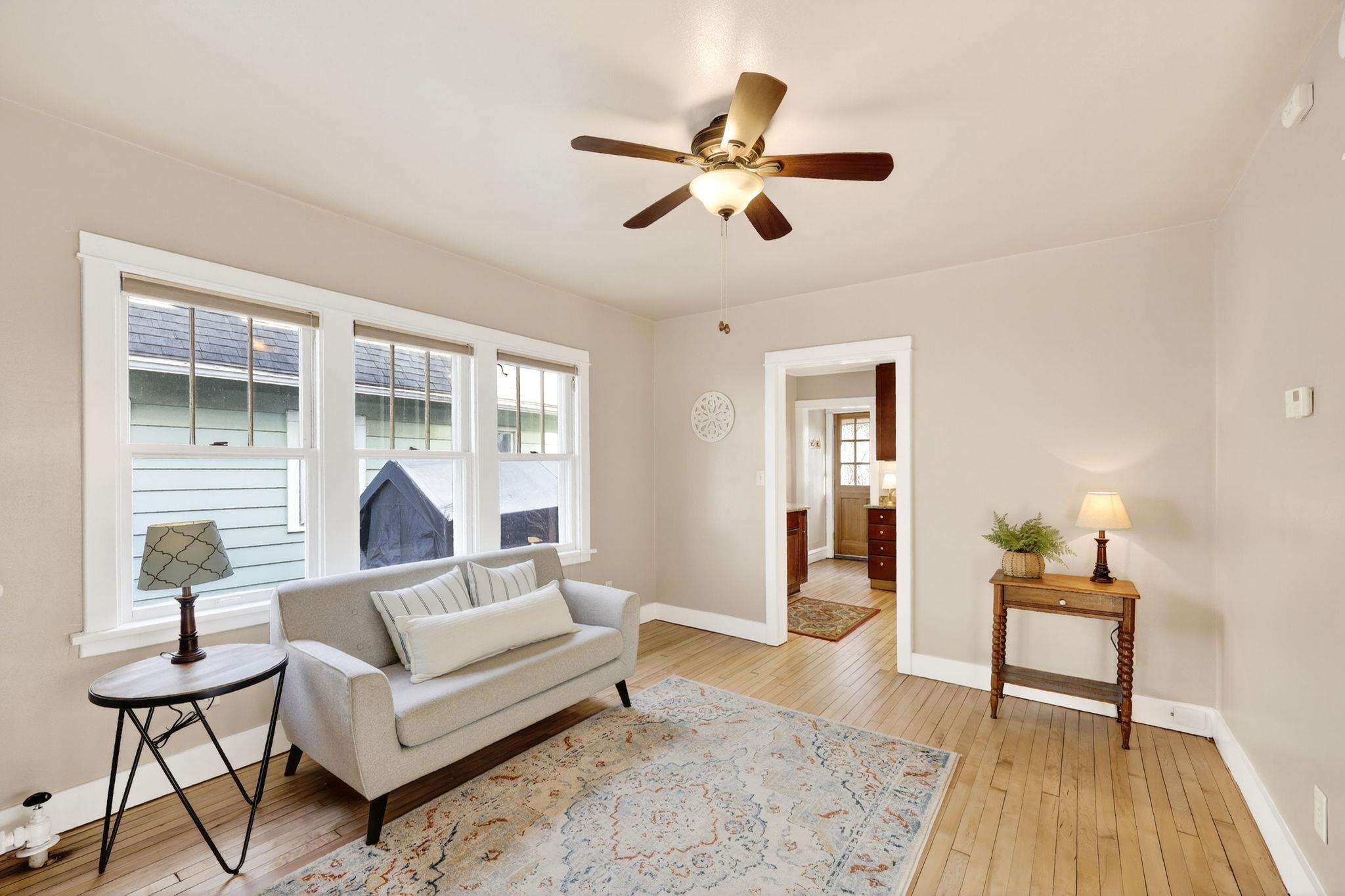 Furnished living room with a gray sofa, hardwood floors, three windows, and a ceiling fan.