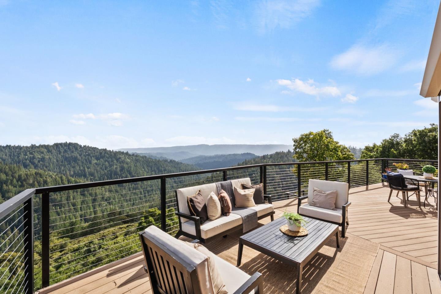 Furnished deck overlooking forested mountain ridges, with cushioned seating and black cable railing.