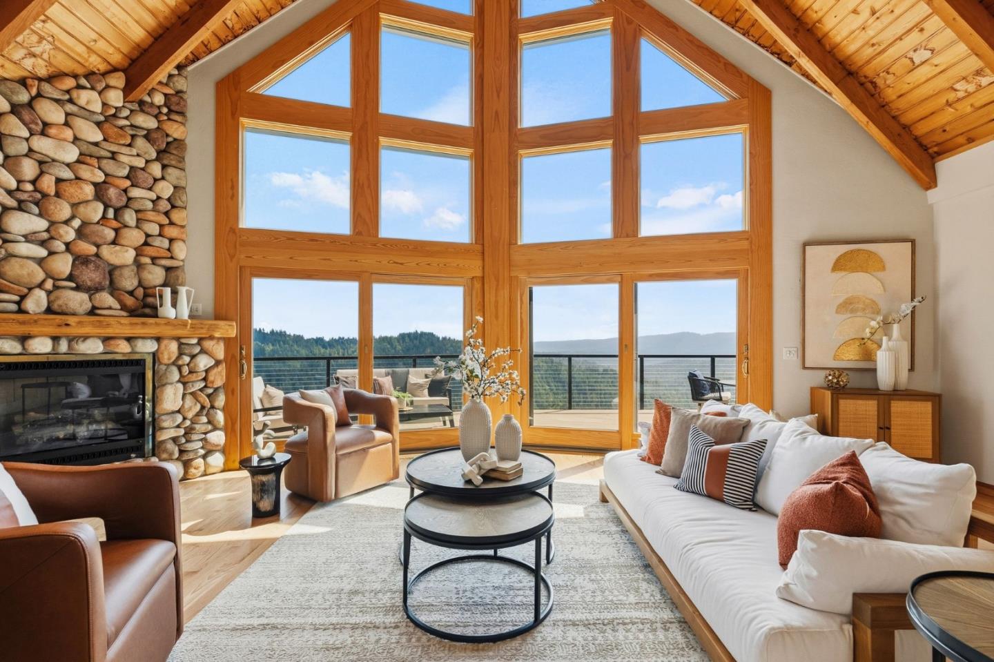 Living room with a stone fireplace, vaulted wood ceiling, and tall windows overlooking wooded hills.