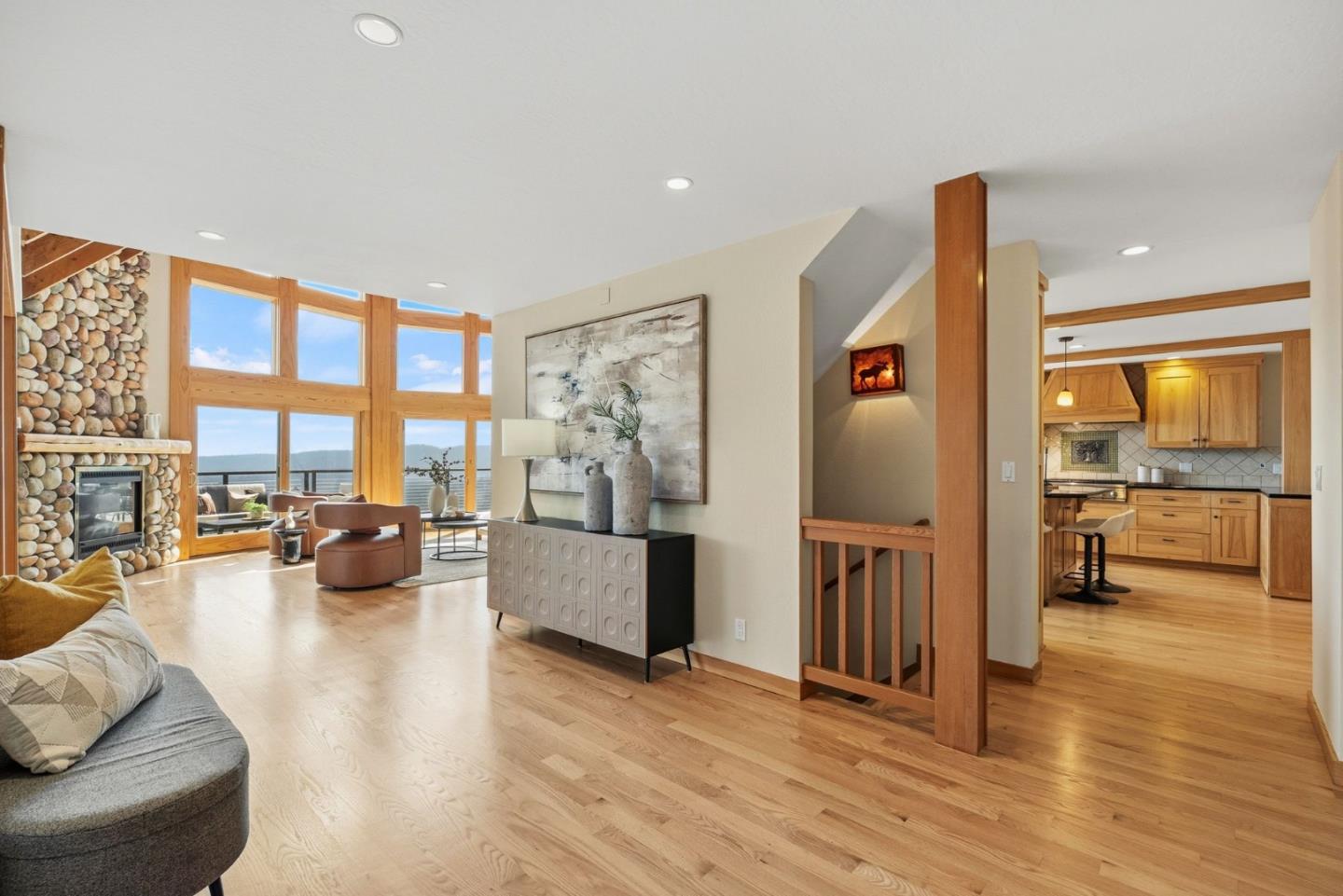 Living room with a stone fireplace, tall windows, and hardwood floors adjoining the kitchen.