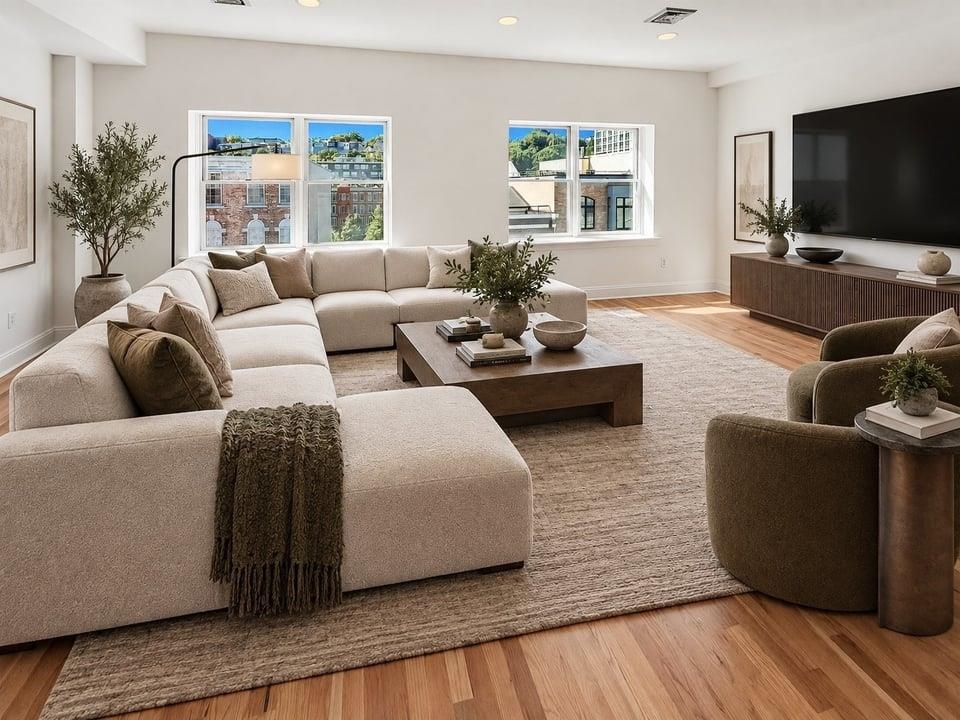 Furnished living room with a cream sectional, wood coffee table, and windows overlooking neighboring buildings.
