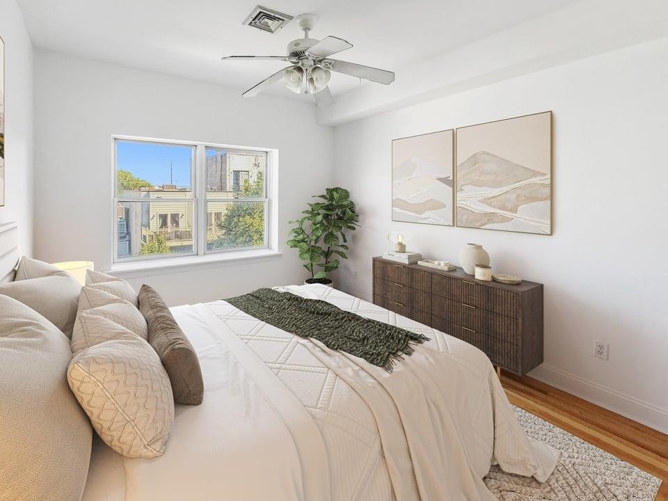 Bedroom with a white-dressed bed, dark wood dresser, ceiling fan, and wide window.