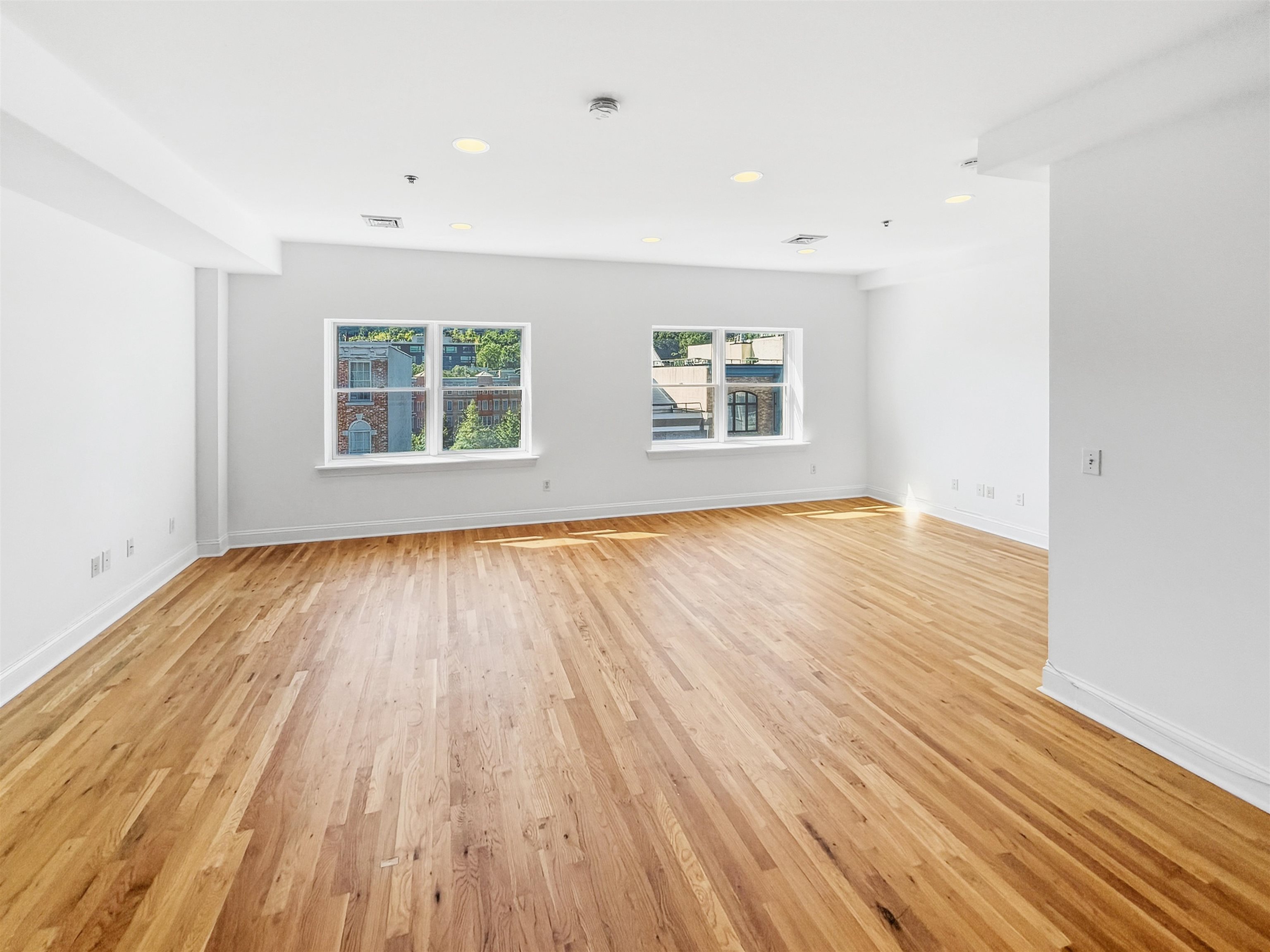 Empty living area with hardwood floors and two windows overlooking nearby buildings.