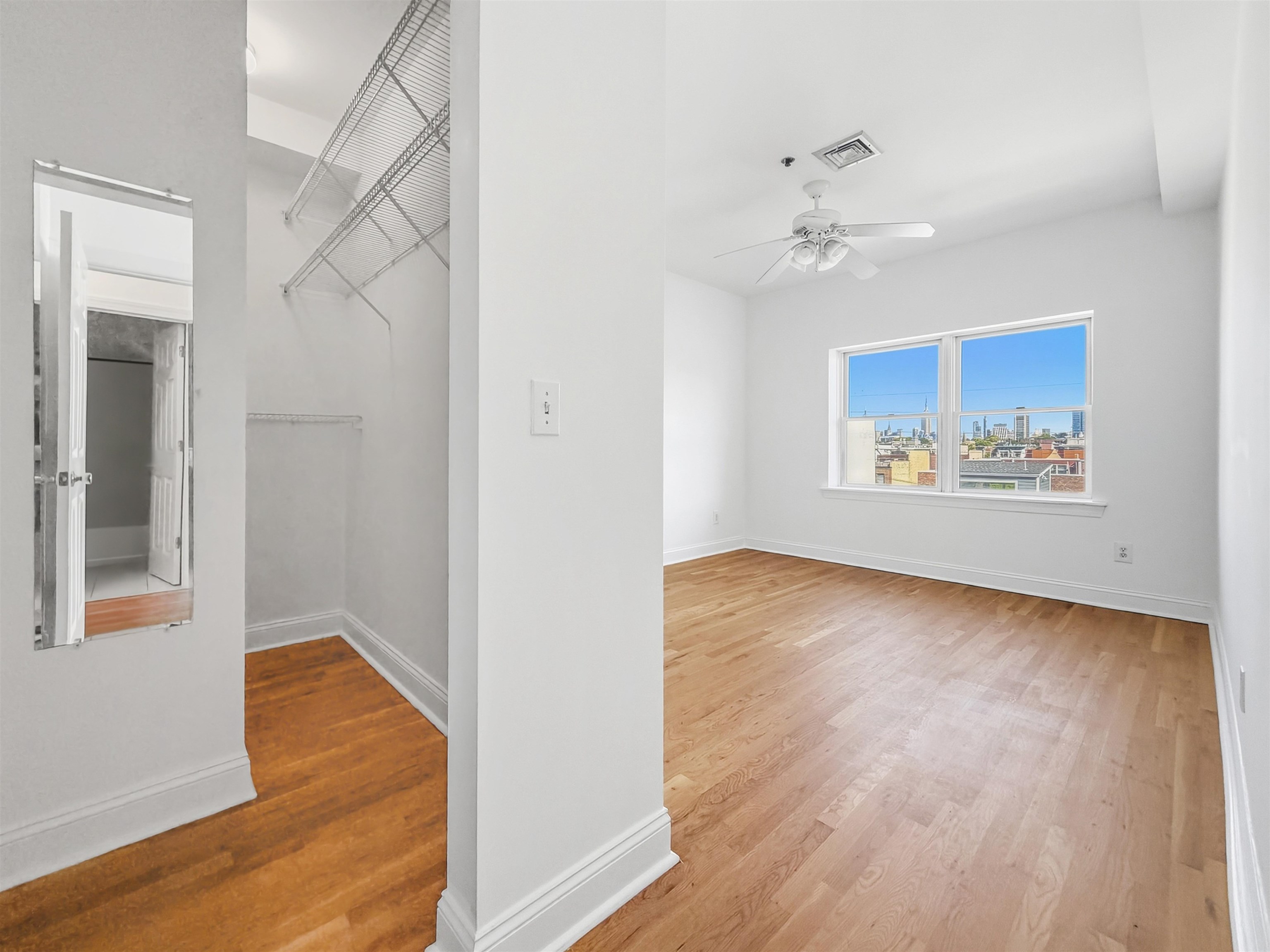 Empty bedroom with hardwood floors, wire-shelved closet, and city skyline view through a double window.