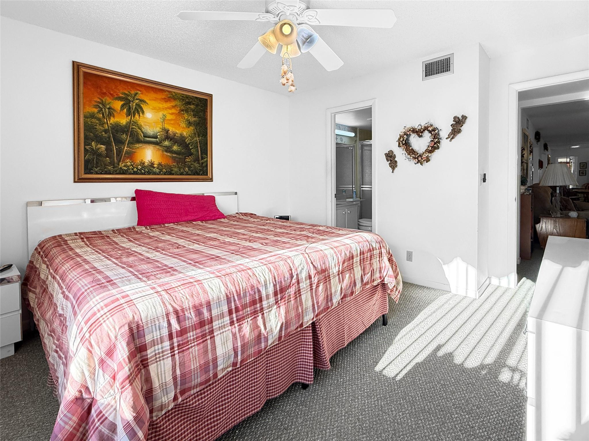 Bedroom with a red plaid bedspread, tropical sunset painting, and adjoining bathroom doorway.