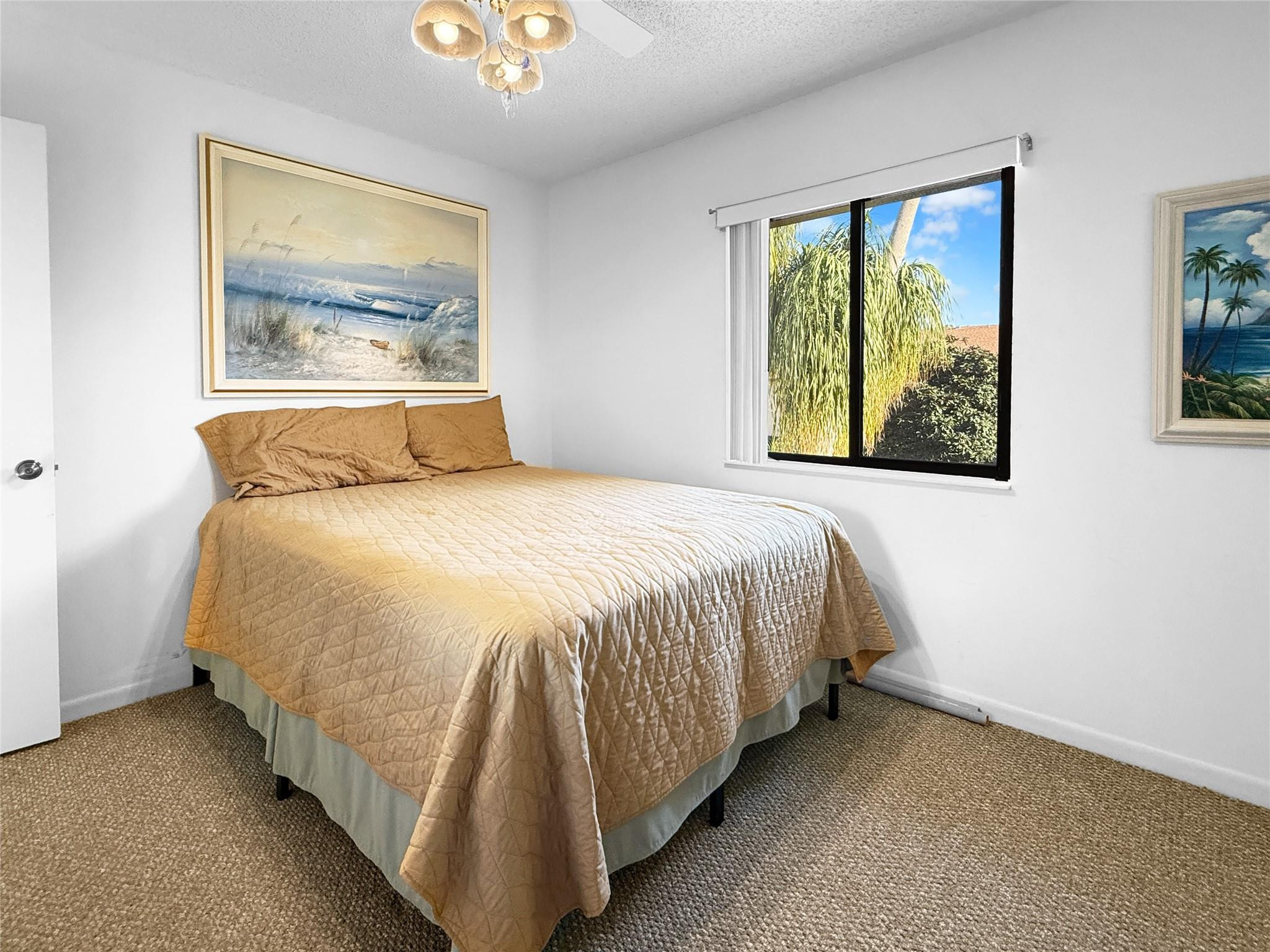 Furnished bedroom with a tan quilted bed and a window overlooking tropical greenery.
