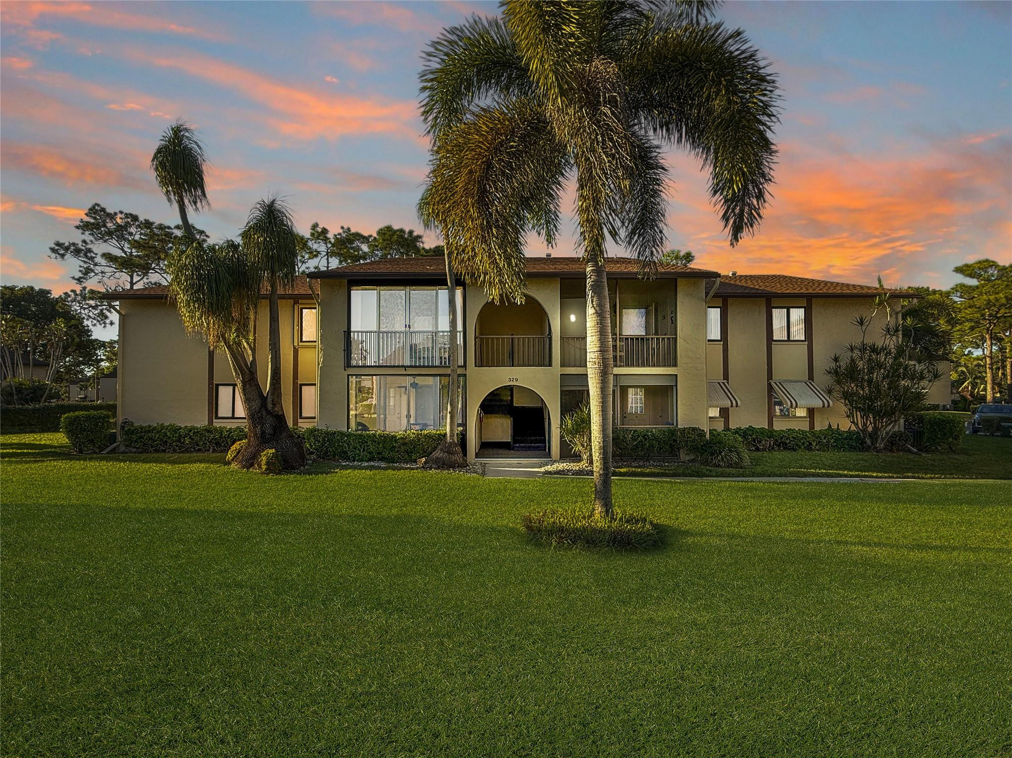 Two-story condominium building with balconies and palm trees beneath a colorful dusk sky.