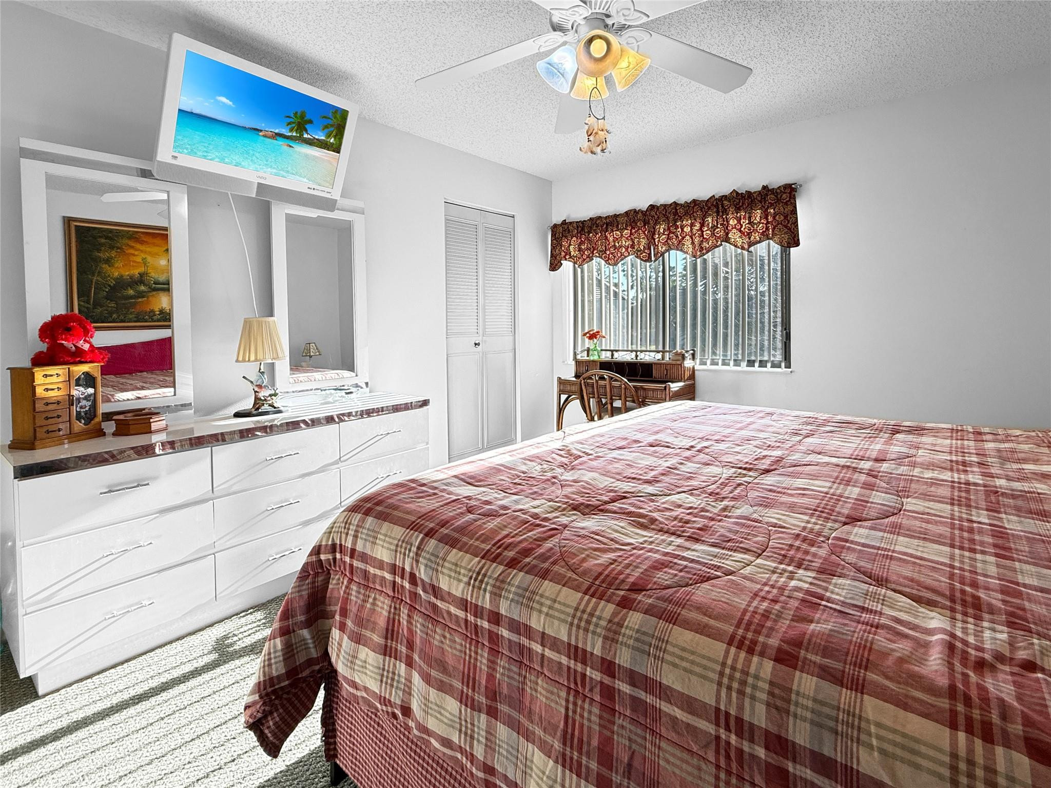 Bedroom with a plaid-covered bed, mirrored dresser, ceiling fan, and window with vertical blinds.