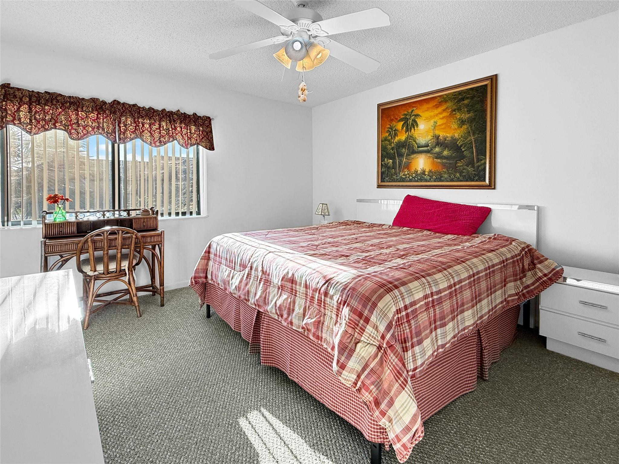 Bedroom with a plaid-covered bed, bright pink pillow, and wooden desk beside a sunlit window.