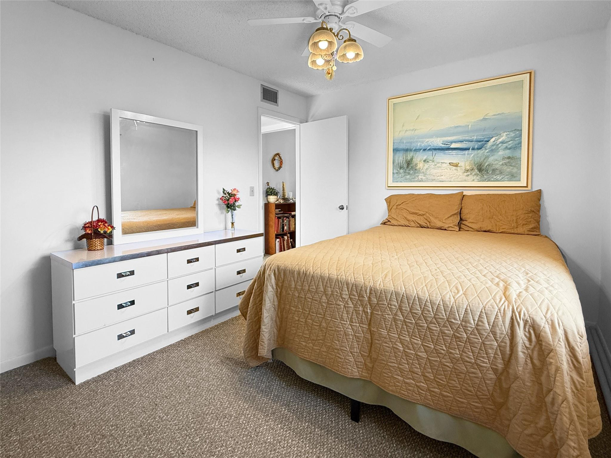 Bedroom with a tan quilted bed, white dresser and mirror, and framed beach artwork.