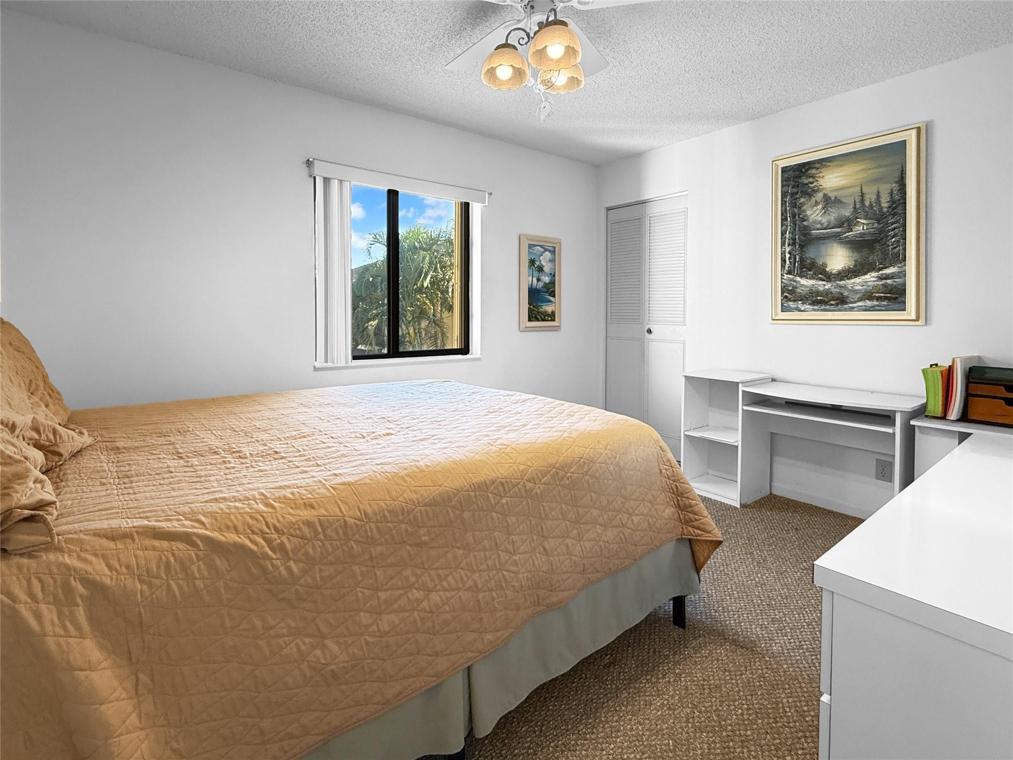 Bedroom with a tan quilted bed, white desk, and window overlooking palm trees.