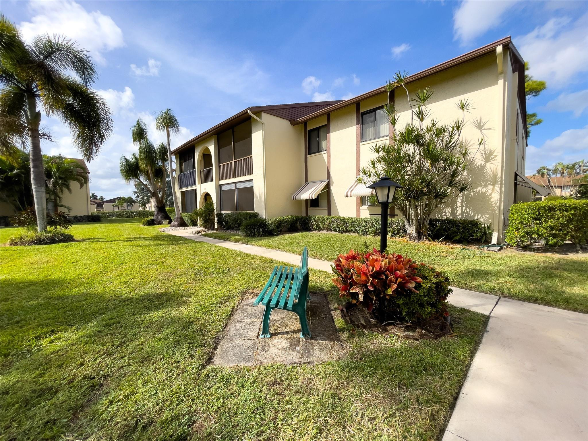 Two-story cream condominium with balconies and awnings beside a landscaped lawn and teal bench.