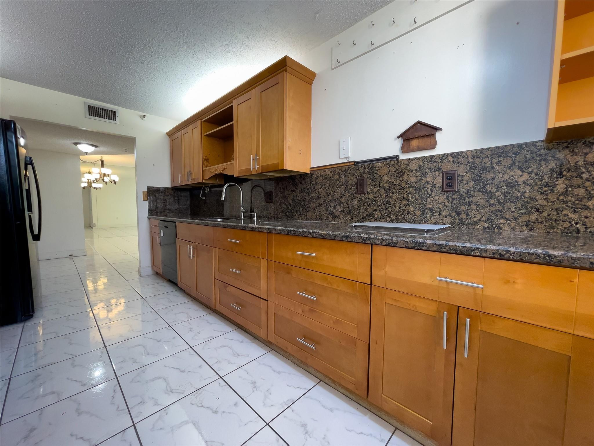 Kitchen with honey-colored cabinets, dark patterned counters, and white marble-look tile flooring.