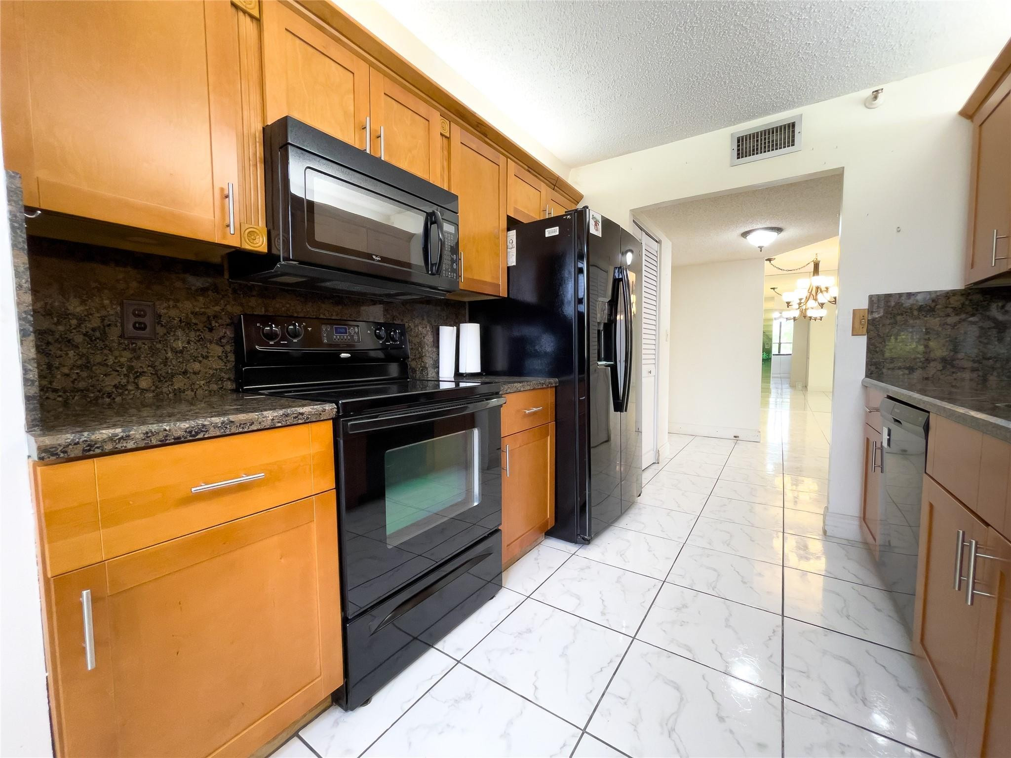 Galley kitchen with honey-colored cabinets, dark granite counters, black appliances, and white tile flooring.