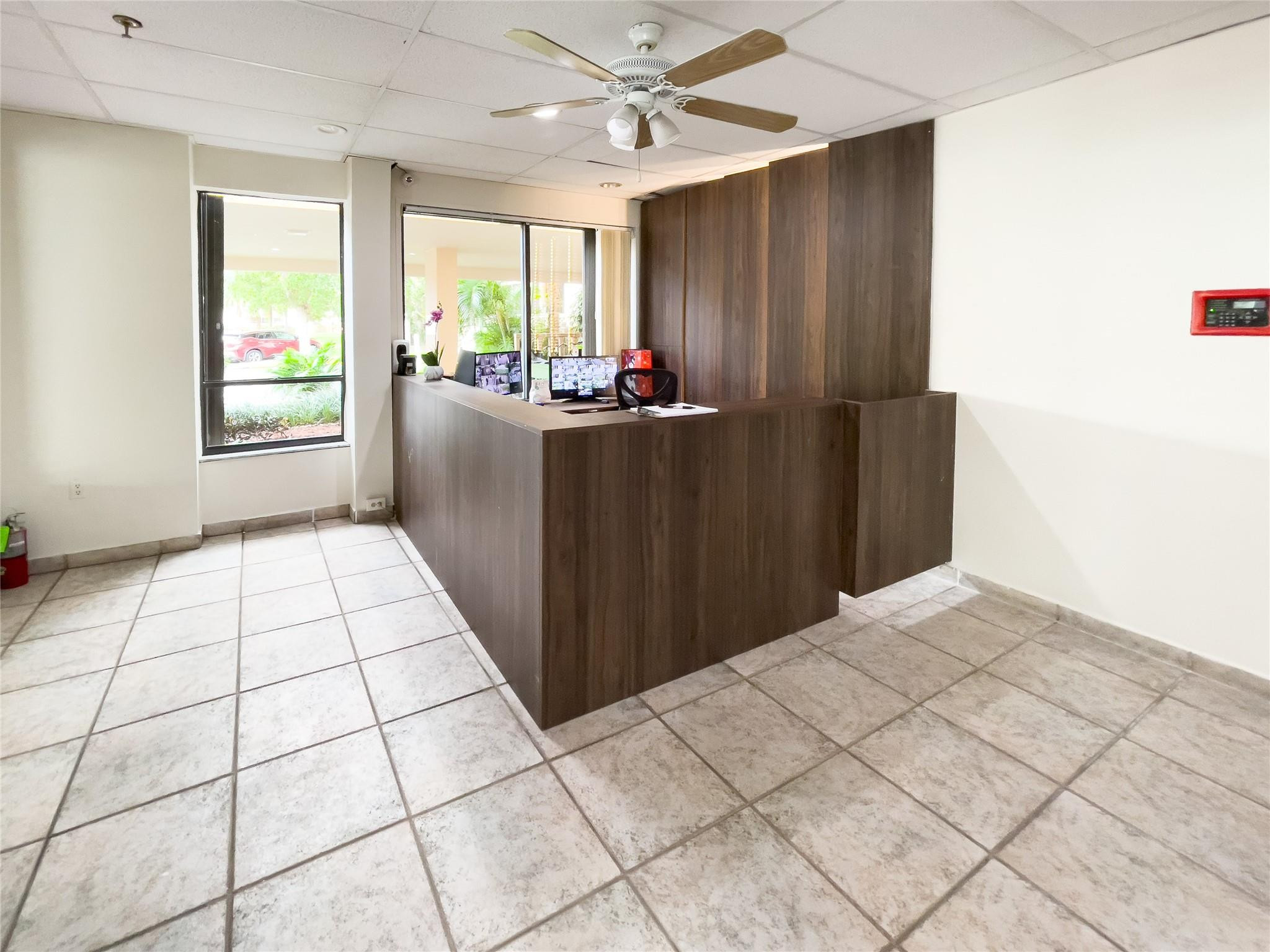 Reception area with a dark wood desk, tiled floor, and windows overlooking greenery.