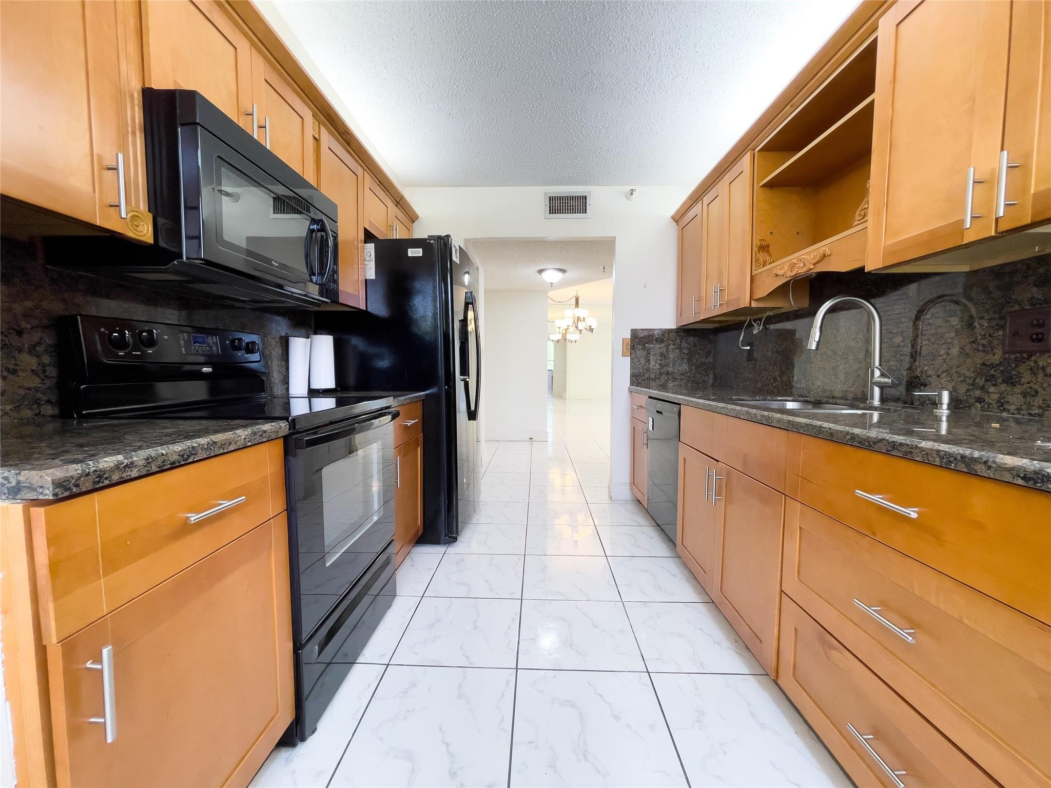 Galley kitchen with honey-colored cabinets, dark countertops, black appliances, and white tile flooring.